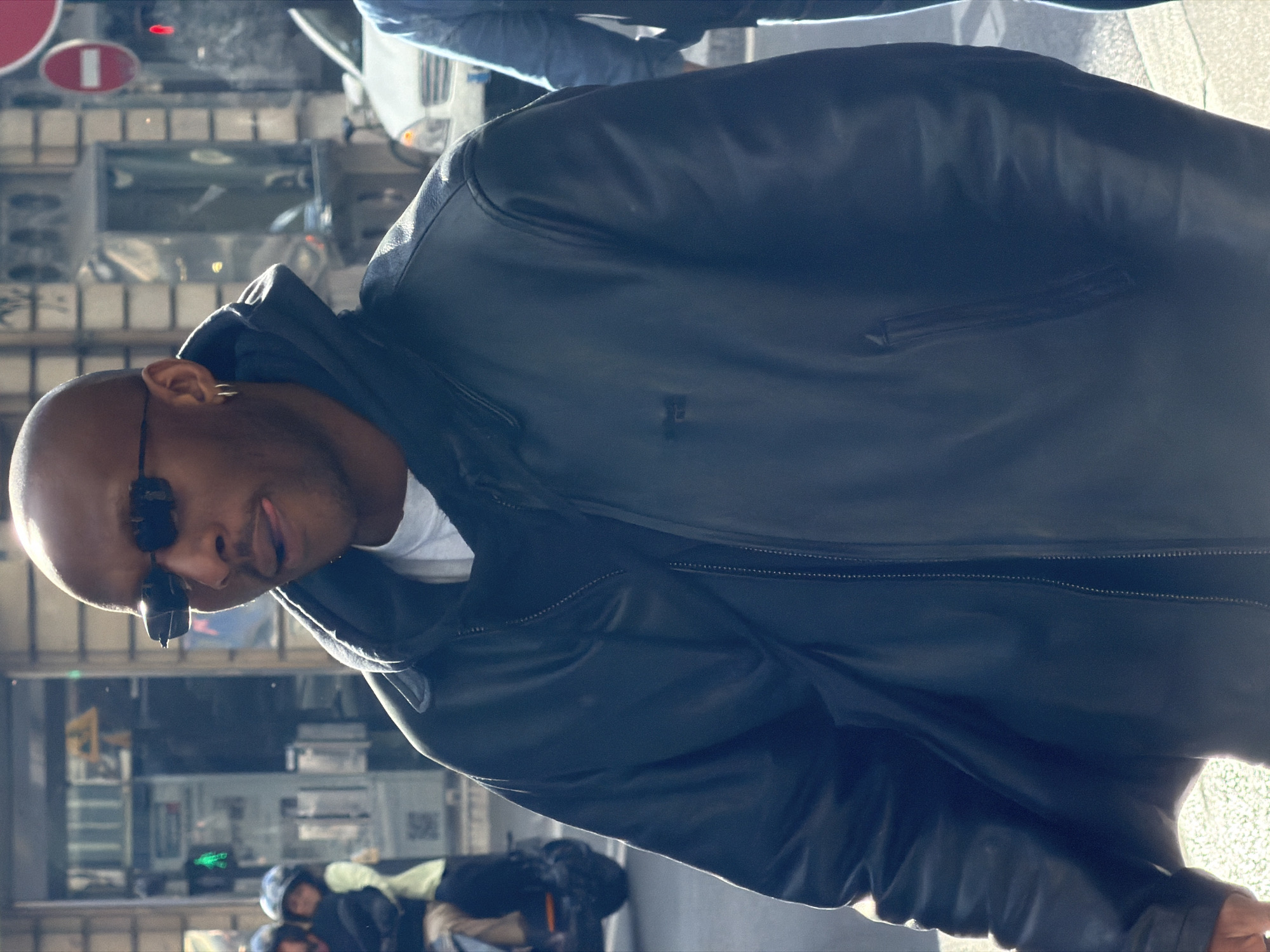 This image captures a man in a tilted perspective, standing outdoors in a sunlit urban environment. He is the primary subject, shown from the chest up, with a shaved head and dark skin. He wears dark, sleek sunglasses and a gold hoop earring in his right ear, with a playful expression that includes his tongue slightly extended. He is dressed in a dark, likely black, leather jacket, worn over a white t-shirt. The jacket features a central zipper and subtle detailing.
The background is dominated by reflections, suggesting the photo was taken near a large window or glass surface. These reflections reveal elements of a busy city street, including indistinct architectural details, other people (some wearing helmets, possibly on scooters or motorcycles), and vehicles. A noticeable red circular traffic sign with a white horizontal bar, commonly indicating "No Entry" in European cities, is clearly visible in the reflection. A faint QR code is also discernible in the background reflections, but its text is illegible.
The bright and clear illumination suggests it is daytime and sunny. The man's confident and relaxed pose, combined with the urban setting and the specific traffic sign, points to a casual street scene in a city like Paris, France, as indicated by the location context.