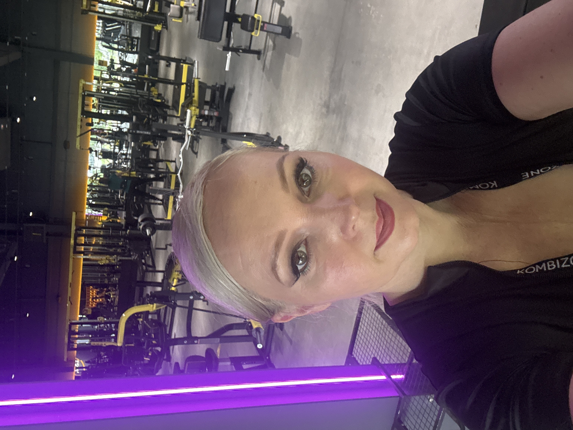 A woman with blonde hair and full makeup is taking a selfie in a brightly lit gym. The gym has many rows of weight machines and exercise equipment, with a prominent purple neon light fixture on the left side of the frame. The woman is wearing a black zip-up top with a logo visible on the neckline. The background shows more gym equipment, including weight racks and benches. There are no visible indicators of the city of Komsomolsk-on-Amur, Russia in the image.