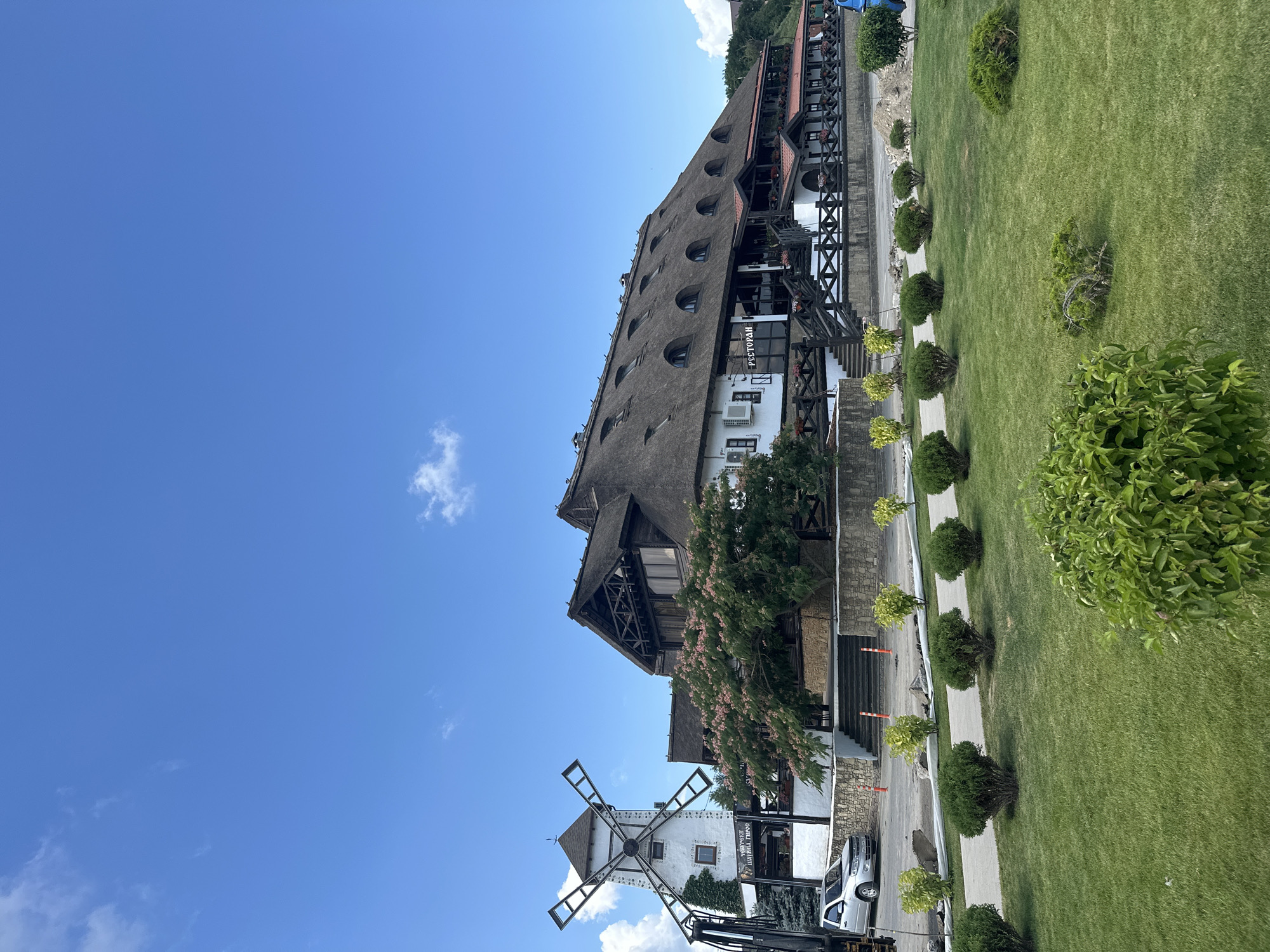 The image shows a building with a thatched roof, a windmill, and a green lawn with bushes. The building appears to be a restaurant, as indicated by the Cyrillic word "РЕСТОРАН" visible on the facade. The windmill is white with four blades. The lawn is well-maintained, and there are several bushes and trees around the property. The sky is blue with some white clouds. The overall impression is that of a pleasant outdoor setting.