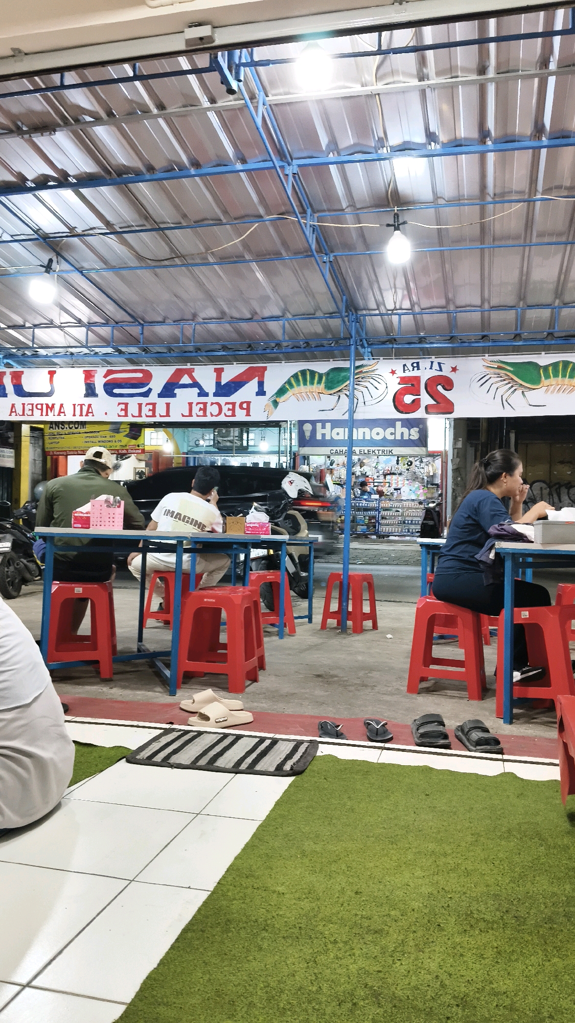 The image depicts an outdoor eating area under a corrugated metal roof supported by blue metal beams. Several tables with blue metal frames and red plastic stools are arranged on a concrete floor. Green artificial turf covers a portion of the ground in the foreground. In the background, there are signs, one of which appears to advertise "NASI PECEL LELE" and "Hannocho's CAHA ELEKTRIK".
Four individuals are visible. In the left half of the image, two men are seated at a table. One man, wearing a green jacket and a beige cap, faces away from the camera. The other man, wearing a white t-shirt with the word "IMAGINE" printed on it, is also seated and facing away. To the right, a woman in a dark blue shirt and pants is seated at another table, facing the right side of the frame and appears to be eating or looking at something in front of her. In the far left foreground, the back of a person wearing white clothing is visible, sitting on what appears to be a green rug.
Various objects are present, including red plastic stools, a blue metal table, food packaging (possibly tissue boxes), and several pairs of footwear (sandals and flip-flops) scattered on the ground. Motorcycles are parked in the background, suggesting a roadside eatery. The lighting in the scene appears to be from overhead bulbs. The environment suggests a casual dining setting, possibly a food stall or a small restaurant. The presence of the sign "Hannocho's CAHA ELEKTRIK" along with the food signage suggests the location might be in a commercial area. The overall impression is of a typical Indonesian street-side eatery.