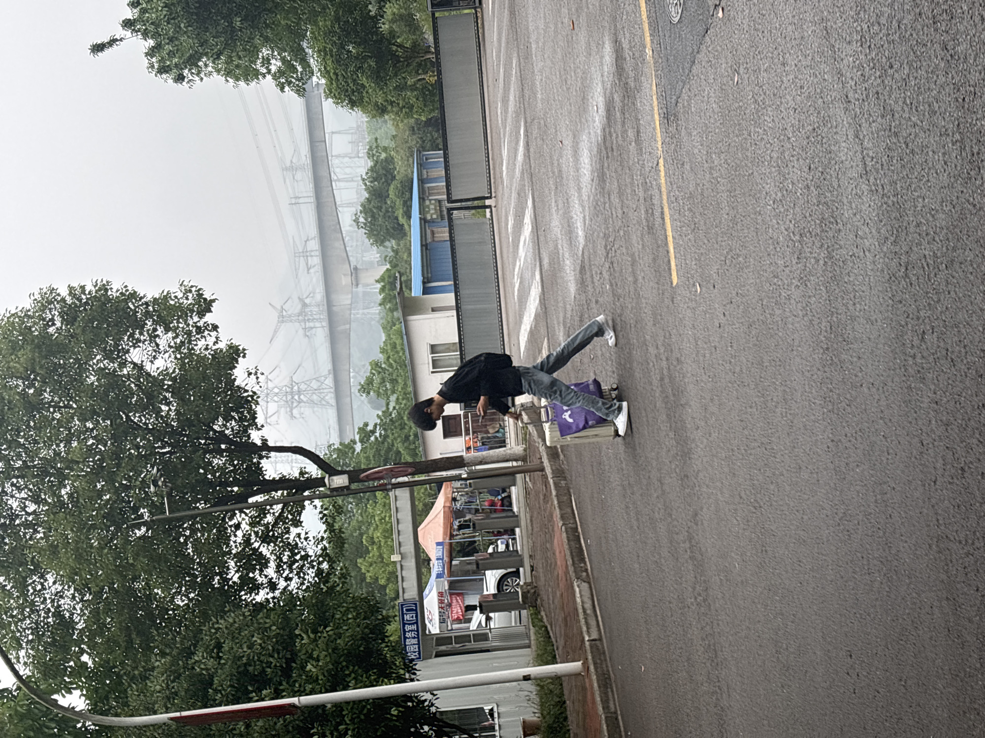 The image shows a street scene in Jiangjin, China. A young man, wearing a dark jacket and jeans, is standing on top of a suitcase on the side of the road. The suitcase is next to a curb along a street with a yellow dividing line. There are buildings and trees in the background, with overhead power lines also visible. There is a sign in Chinese, and an awning in the background as well as cars parked in the background.