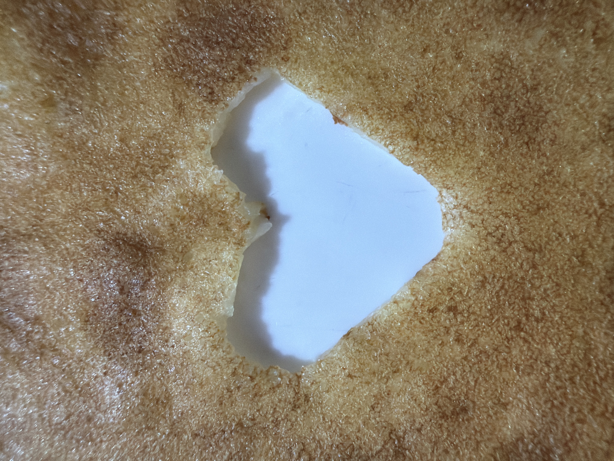 A close-up shot shows a thin, golden-brown layer with an irregular hole exposing a white surface beneath. The layer has a textured, almost granular appearance with slight variations in color, ranging from light tan to darker brown patches. The hole's edges are slightly uneven and frayed. The lighting appears to be artificial, creating a somewhat glossy sheen on the surface. Location information for Chaikovsky, Russia, is not discernible from the visual data in this image.