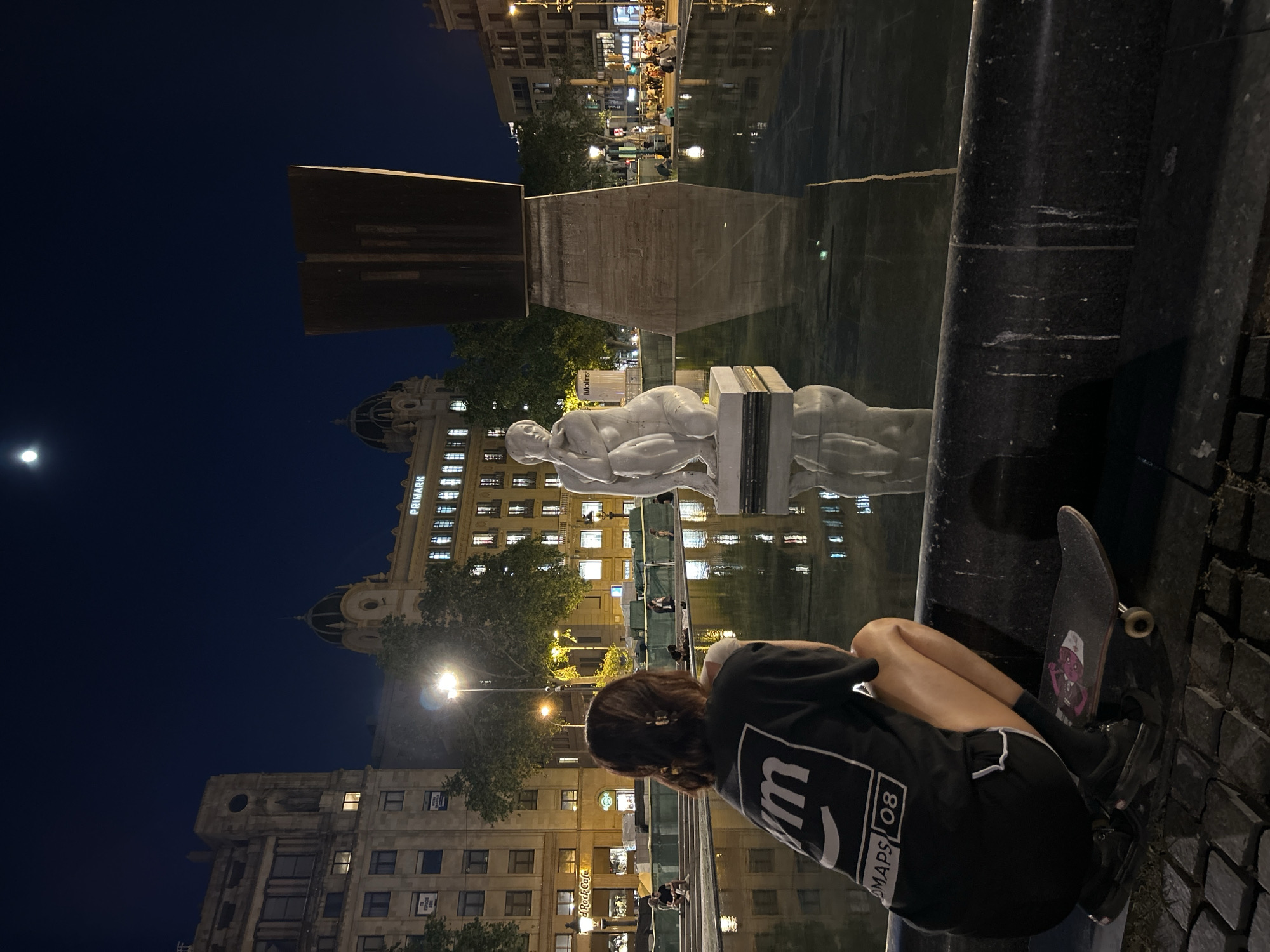 A person is sitting near a fountain at night in Barcelona, Spain. The person is wearing a black t-shirt, black shorts, and black shoes, and is sitting with their knees drawn up to their chest. A skateboard is next to the person. The fountain has a statue of a reclining figure in the water, and a modern, rectangular sculpture stands behind it. Buildings with lit windows are visible in the background, including a building with a dome and a sign for "PRIMARK." The night sky is dark, with a moon visible. The reflection of the buildings, sculpture, and statue is visible in the water.