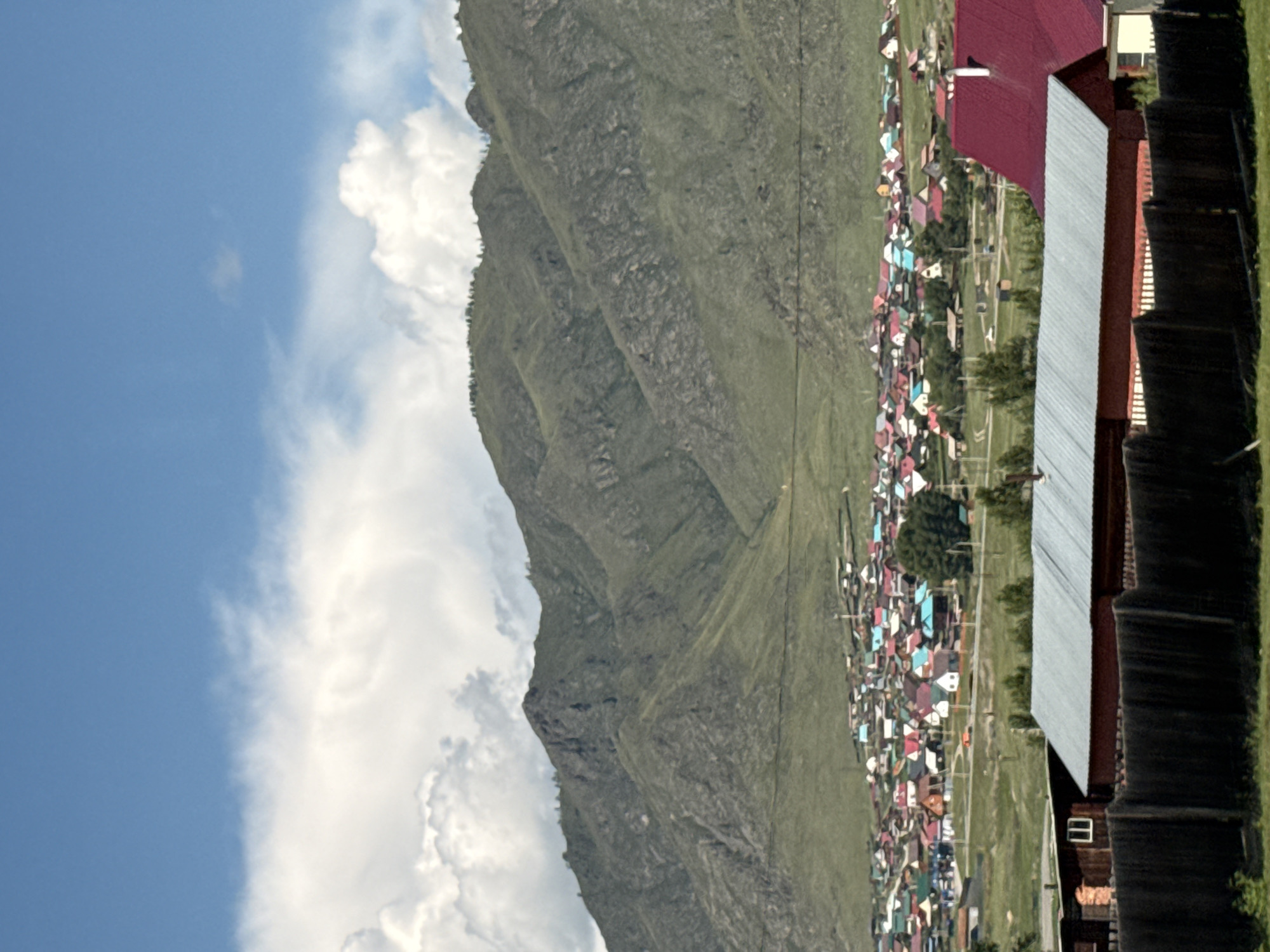 This image depicts a rural village nestled at the foot of a vast mountain range. The village features numerous small houses with brightly colored roofs in shades of red, blue, green, and white, creating a vibrant mosaic against the green landscape. A road winds through the village, with a few vehicles visible. In the foreground, a dark wooden fence runs across the bottom of the frame, partially obscuring two buildings. One building on the left is made of dark wood with a small window, and the other on the right has a red metal roof. The mountains in the background are covered in sparse vegetation, with rocky outcrops and deep ravines. The sky is a clear blue with scattered white clouds. The scene suggests a peaceful, remote settlement in a mountainous region. This scene is likely located in or near the city of Onguday, Russia, a region known for its mountainous terrain and traditional villages.