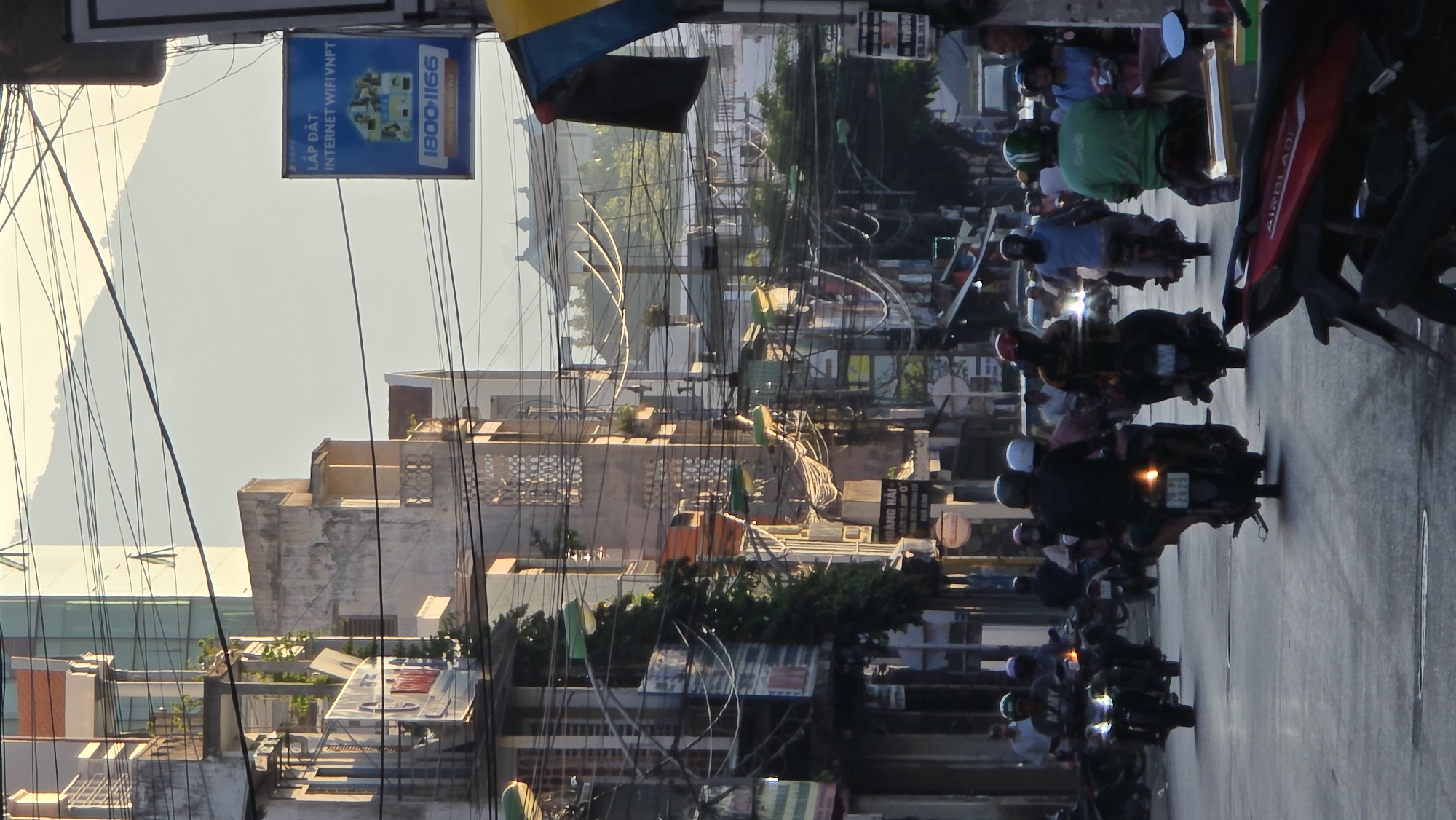 The image shows a street scene in Nha Trang, Vietnam, with a line of motorbikes traveling away from the camera. Several buildings are along the sides of the street. Overhead wires are present in the scene. There are commercial signs in Vietnamese on the buildings. The sky is pale.