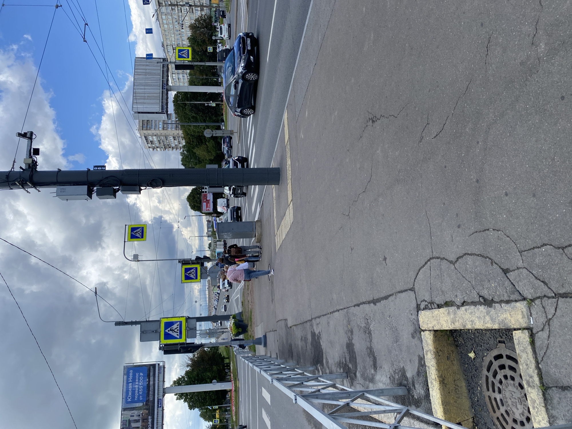 The image depicts a street scene in Saint Petersburg, Russia, specifically at a pedestrian crossing. In the foreground, a grey asphalt sidewalk is visible with cracks and a metal railing. To the left, a manhole cover is partially visible. Across the street, traffic lights and pedestrian crossing signs are mounted on poles. Several cars are on the road, including a dark sedan in the right lane. A few pedestrians are walking on the sidewalk near the crossing. In the background, apartment buildings and trees line the street, under a cloudy sky. A large billboard with Russian text is visible on the left.
