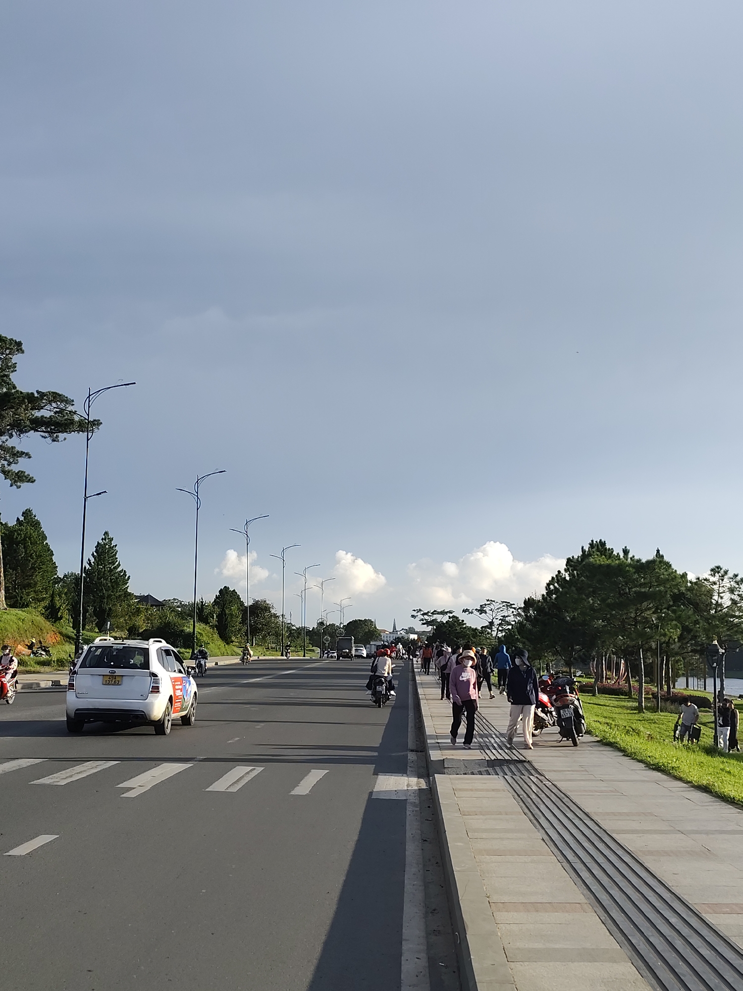 This image depicts a street scene in Da Lat, Vietnam, likely during daylight hours given the illumination and shadow patterns. The foreground features a wide asphalt road with clear white lane markings and a pedestrian crosswalk. A white SUV is driving away from the camera in the left lane. The right side of the road is a paved sidewalk with textured tactile paving strips. A line of people are walking on the sidewalk, some in casual attire, and a few are wearing face masks. Several motorcycles are parked or being ridden on the road and sidewalk.
Adjacent to the sidewalk is a grassy embankment with numerous trees, including tall pine trees characteristic of the Da Lat region. In the background, more trees and vegetation are visible, along with a glimpse of distant buildings. The sky is a pale blue with scattered white clouds. The overall atmosphere suggests a leisurely day in a scenic urban environment.
