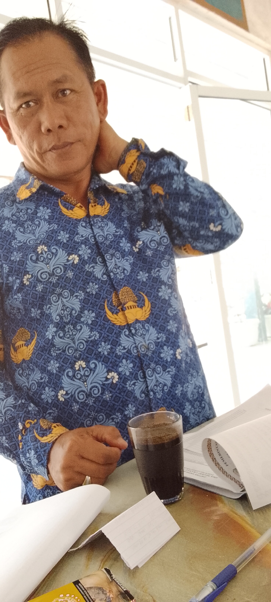 A middle-aged Indonesian man with short, dark hair is pictured from the chest up, indoors. He is wearing a blue batik shirt with yellow emblems and a floral pattern. He is touching the back of his neck with his right hand. In front of him, on a table, is a glass of dark liquid, possibly coffee, some crumpled papers, and an open document. The background shows a window with white frames, suggesting a bright, possibly sunny day. The man's expression is neutral.