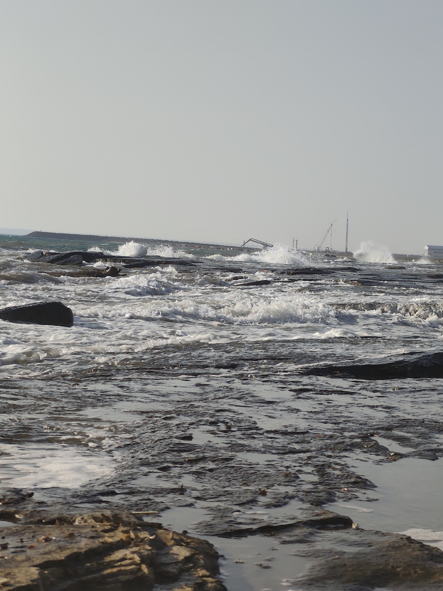 This is a photograph of a rocky coastline with choppy water. The sky is overcast and pale. In the distance, there are construction cranes and buildings, suggesting an industrial or port area. Waves are crashing against the rocks, creating white foam. The foreground consists of wet, dark rocks with some shallow pools of water reflecting the sky. The overall impression is one of a somewhat industrial and weathered coastal environment. The location is specified as the city of Saiyn Şapağatov, Kazakhstan. There are no people or animals visible in the image.