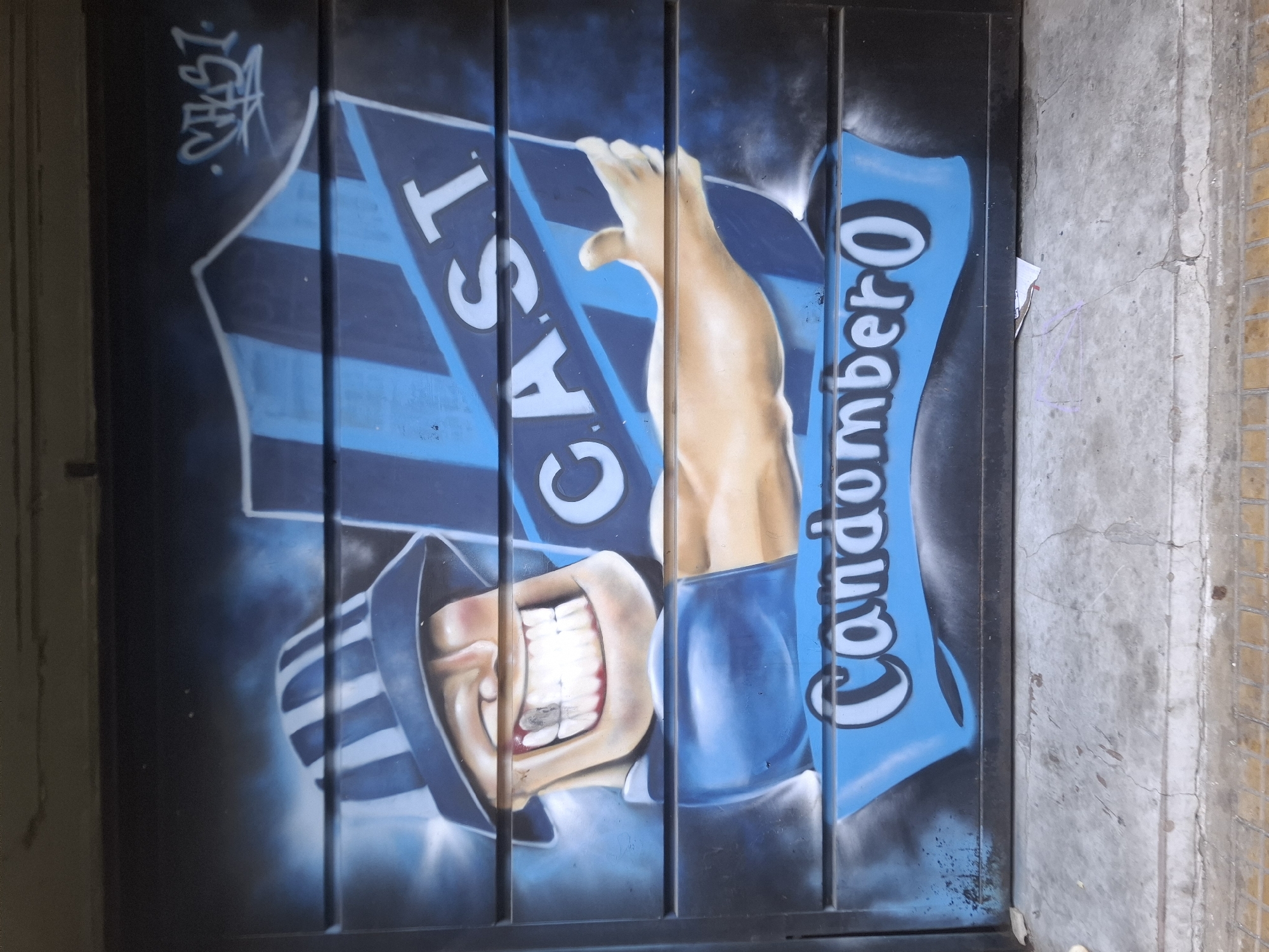 The image shows graffiti art on a corrugated metal door. The artwork depicts a cartoon figure wearing a striped hat and displaying a wide grin, a human arm and hand, a striped banner, and the word "CAST". To the figure's right, another banner is painted with the word "Condombero". The backdrop is a mix of blue and black spray paint. The door is situated between a grey concrete wall on the right and an indistinct wall on the left. The location is Buenos Aires, Argentina.