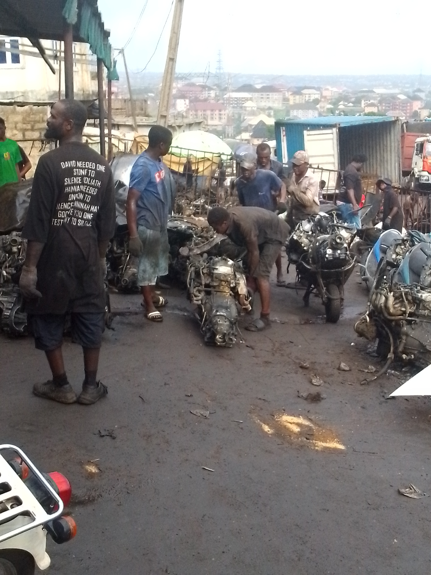 The image captures an outdoor motorcycle engine repair or salvage yard in Onitsha, Nigeria. Approximately ten men are visible, most engaged in handling or inspecting numerous disassembled or partially assembled motorcycle engines scattered across the dark, worn ground. The ground appears to be a mix of dirt and possibly oil, with various debris.
In the foreground left, the rear section of a motorcycle with a white fender and red taillight is visible. Further back, men are bending over engines, suggesting active work or inspection. The sky is overcast, creating a soft, diffuse light that indicates either early morning, late afternoon, or a heavily cloudy day.
The background reveals a bustling urban landscape with numerous buildings stretching into the distance. Shipping containers, including one blue and one white, are prominent, along with a truck with a red cab, hinting at a commercial or industrial zone. Overhead, rudimentary structures with green tarp covers provide shelter or shade.
A man in the foreground, seen from behind, wears a black t-shirt with white text. The visible text, despite some misspellings, conveys a motivational or religious message: "DAVID NEEDED ONE STONF TO SILENCE FOLIATH HANNA NEEDED ONTON TO ILENCENNINAH MA GOVE YOU ONE TESTINY TO SMAGE." The scene conveys a busy, industrious atmosphere characteristic of a vibrant local market or workshop.