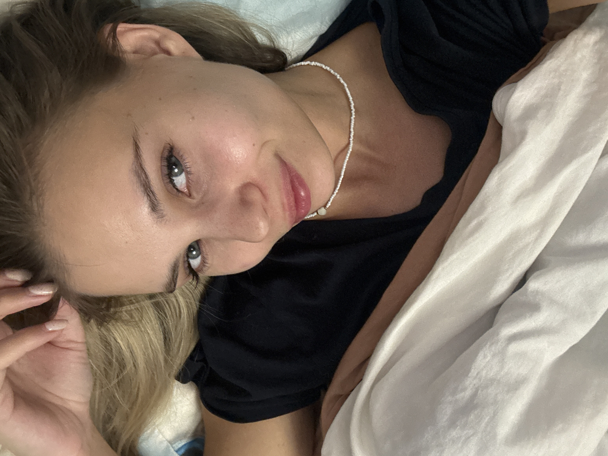 The image displays a young woman lying down indoors, likely in a bed, captured from a slightly elevated and angled perspective. Her head is tilted to the left, and she gazes directly at the viewer with a calm and gentle expression, featuring a slight smile. She has light blonde or brown hair, fair skin, and striking blue-grey eyes. She is wearing a dark, possibly black, top with a ribbed texture. A delicate white beaded necklace with a small, light-colored pendant adorns her neck. Her left hand, with light-colored manicured nails, is visible near her hair, close to her temple.
The setting appears to be a bedroom, with various fabrics creating the background. A light blue fabric is visible behind her head, suggesting a pillow or sheet. To her right, a voluminous white duvet or sheet is present, along with a section of light brown or tan fabric. The lighting is soft and even, characteristic of an indoor environment without strong natural sunlight or artificial light sources. The overall mood is intimate and relaxed. No specific time of day, weather conditions, or textual elements are discernible from the image. This scene is located in Chaikovsky, Russia.