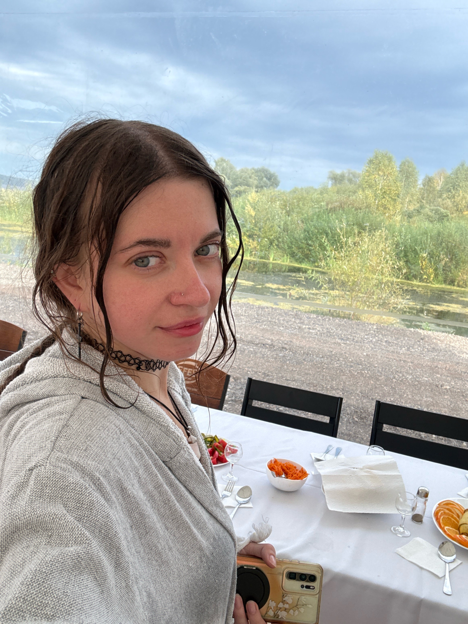 A young woman with wet, dark brown hair pulled into a braid and loose strands framing her face is looking directly at the camera. She has light blue eyes, a nose piercing, and is wearing a grey hooded sweatshirt and a black choker necklace. She is holding a gold-colored phone with a floral case in her right hand.
The woman is seated at a white-clothed table set for a meal. There are plates with food, cutlery, glasses, and napkins on the table. In the background, through a clear barrier, is a body of water with lush green vegetation and trees under a cloudy sky. The setting appears to be an outdoor dining area, possibly a restaurant or cafe. The location is identified as Пакли, Russia.