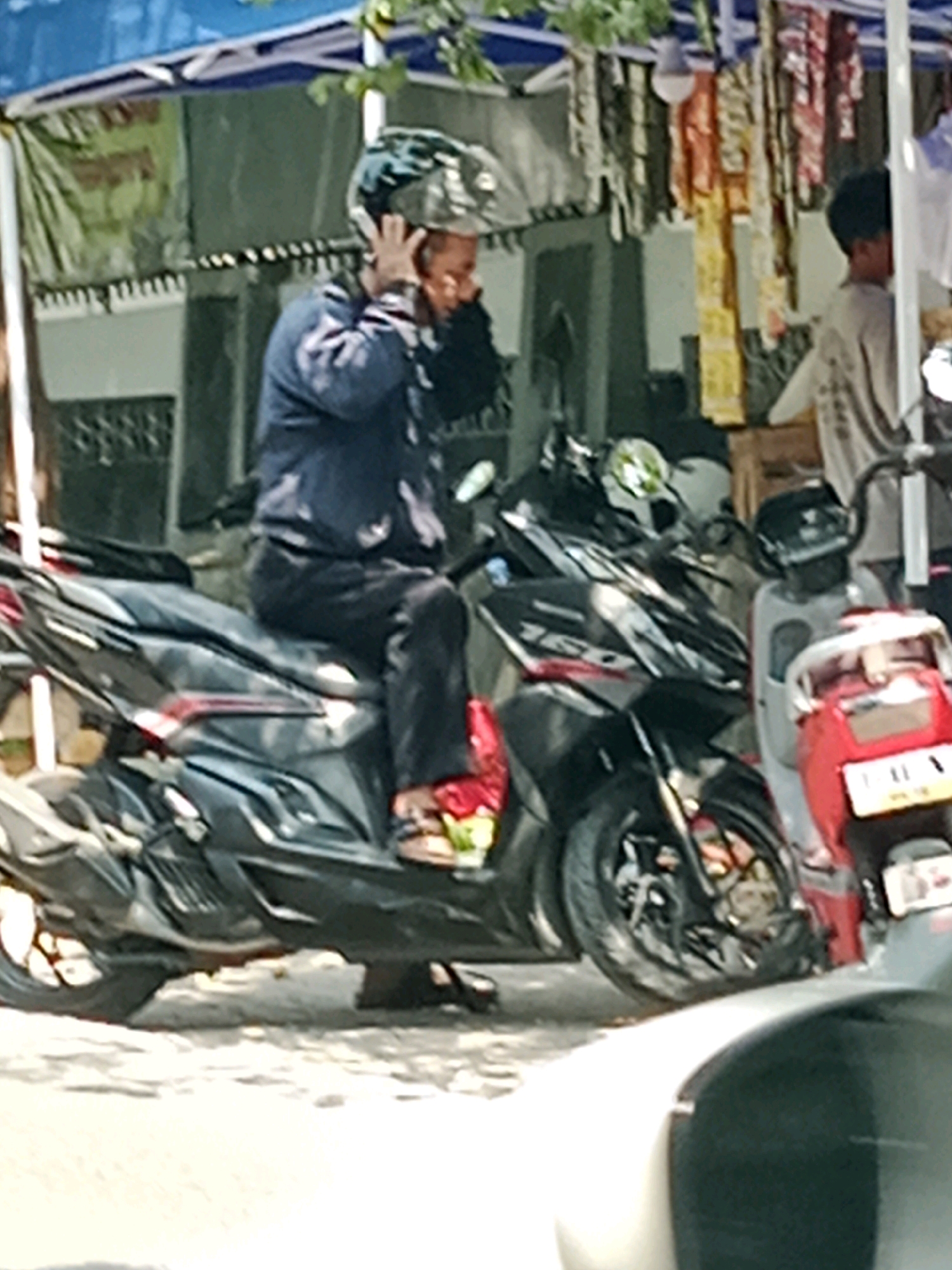 In Banaran, Indonesia, a person wearing a helmet and a blue jacket sits on a black motorcycle. The person is holding their hands to the helmet. In the background, there are buildings, tents, and hanging fabrics, suggesting a market or street vendor area. Another motorcycle is partially visible to the right. The scene appears to be outdoors during the daytime.