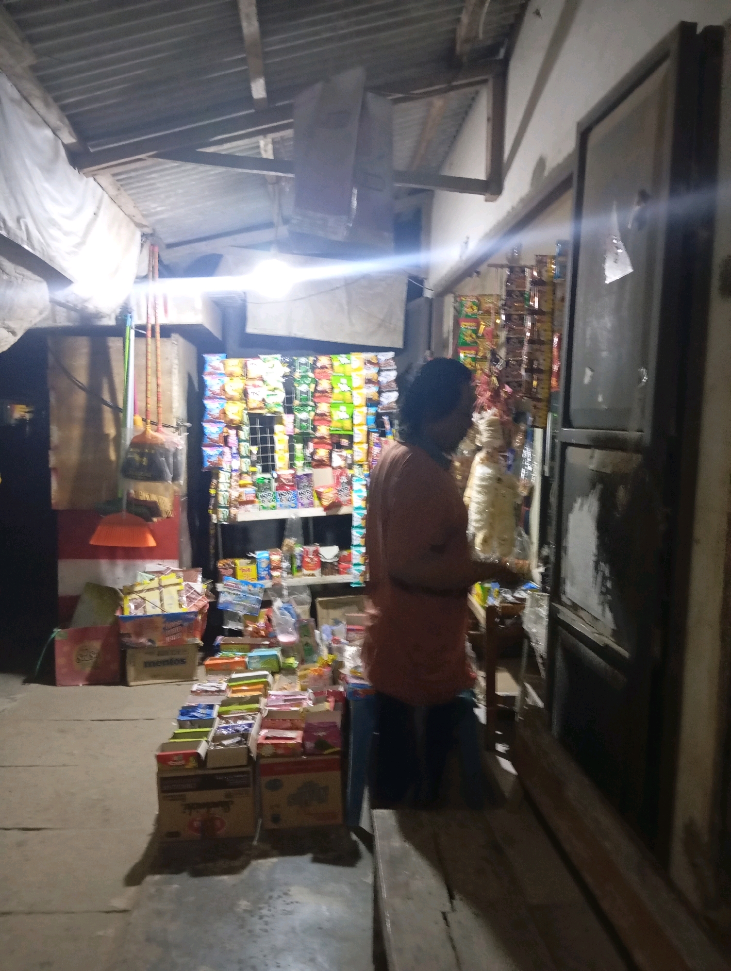 The image depicts a small store, likely a warung, in Leman Benawi, Indonesia, at night. A woman wearing an orange shirt and dark pants stands behind a counter filled with various goods, including snacks, drinks, and other small items. Behind her, shelves are stacked with colorful packaged snacks and products. To the left of the counter, brooms and other cleaning supplies are visible. The store appears to be open-air or semi-open, with a corrugated metal roof supported by wooden beams. The ground is paved with concrete, and a wooden bench is positioned in front of the counter. The lighting suggests it's evening or night, with a bright light source illuminating the area.