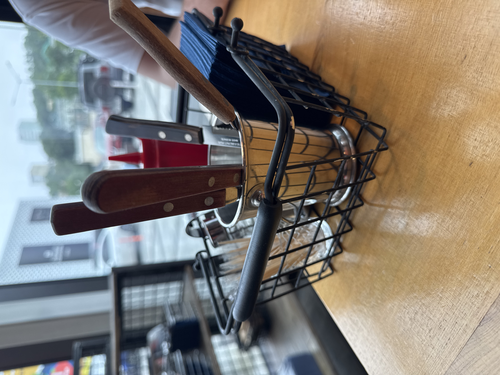 The image depicts a close-up view of a black wire basket containing cutlery, blue napkins, and a metal holder filled with knives. The basket is placed on a wooden table, with a blurry background showing a street scene through a window. The scene appears to be indoors, likely a restaurant or cafe, given the presence of cutlery and napkins. Outside the window, cars and buildings are visible, suggesting an urban environment. The overall lighting is dim, with a focus on the objects in the foreground. The image is taken from a slightly elevated perspective, looking down at the basket. There are no visible people in the foreground, though a person's arm and shoulder are visible in the upper right corner of the image, wearing a white t-shirt. The environment appears to be a public space, possibly a restaurant with a view of a street in Vladivostok, Russia.