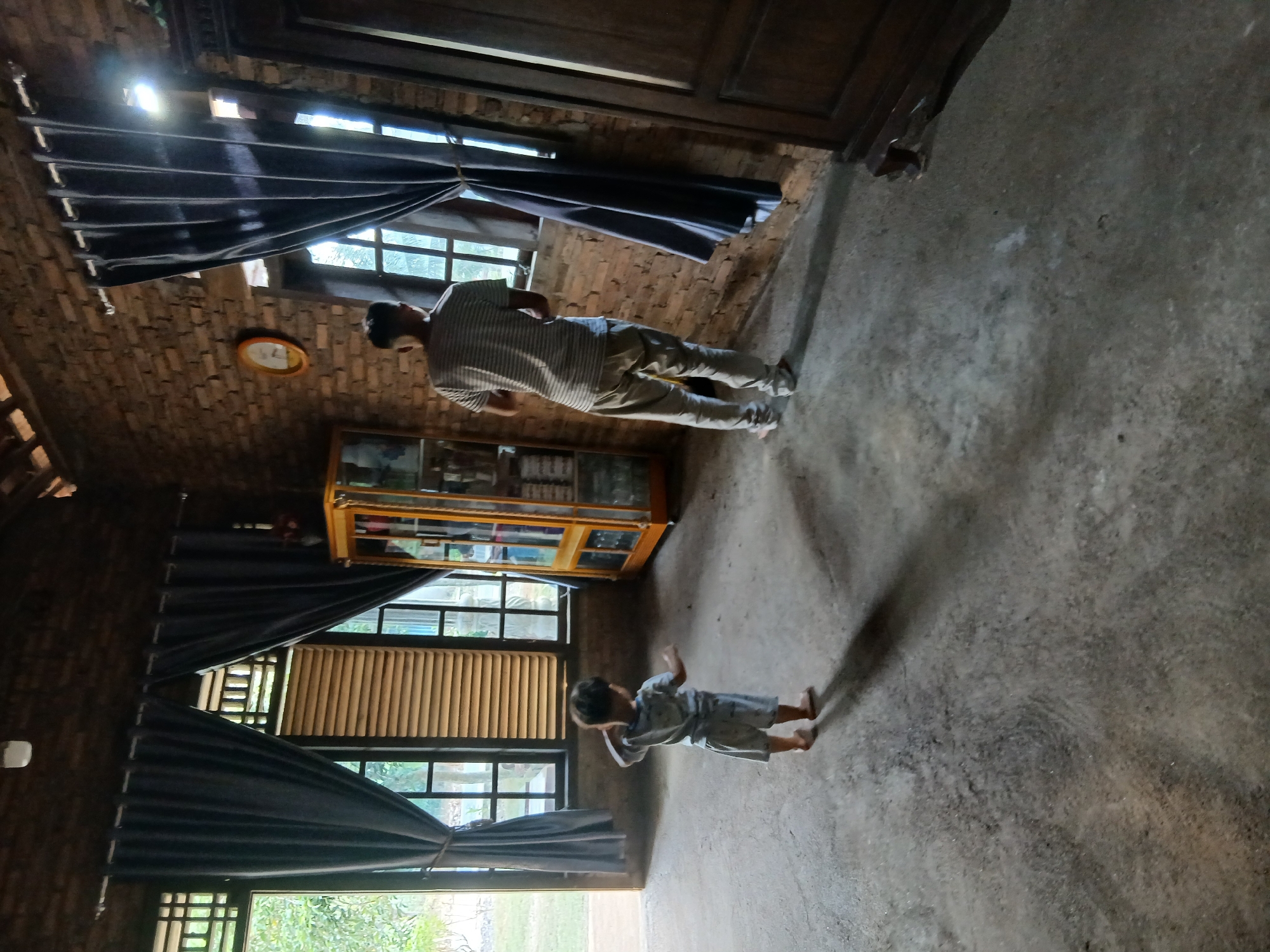 An interior scene is captured within a room constructed of brick walls and a wooden beamed ceiling. The room is illuminated by natural light filtering through windows, casting long shadows on the concrete floor. On the left side of the frame, a young child, appearing to be a toddler, stands with their back to the camera, wearing a light-colored t-shirt and shorts, and is barefoot. Further into the room, an adult male, seen from behind, is wearing a striped t-shirt and light-colored pants. He is standing near a window, with his back partially turned. In the background, a tall, glass-doored cabinet with a golden frame contains various items. To the right, a large, dark wooden cabinet with carved details is partially visible. The environment suggests a rustic or traditional dwelling, possibly in Kalirejo, Indonesia, given the architectural style and climate implied by the open windows and natural light.