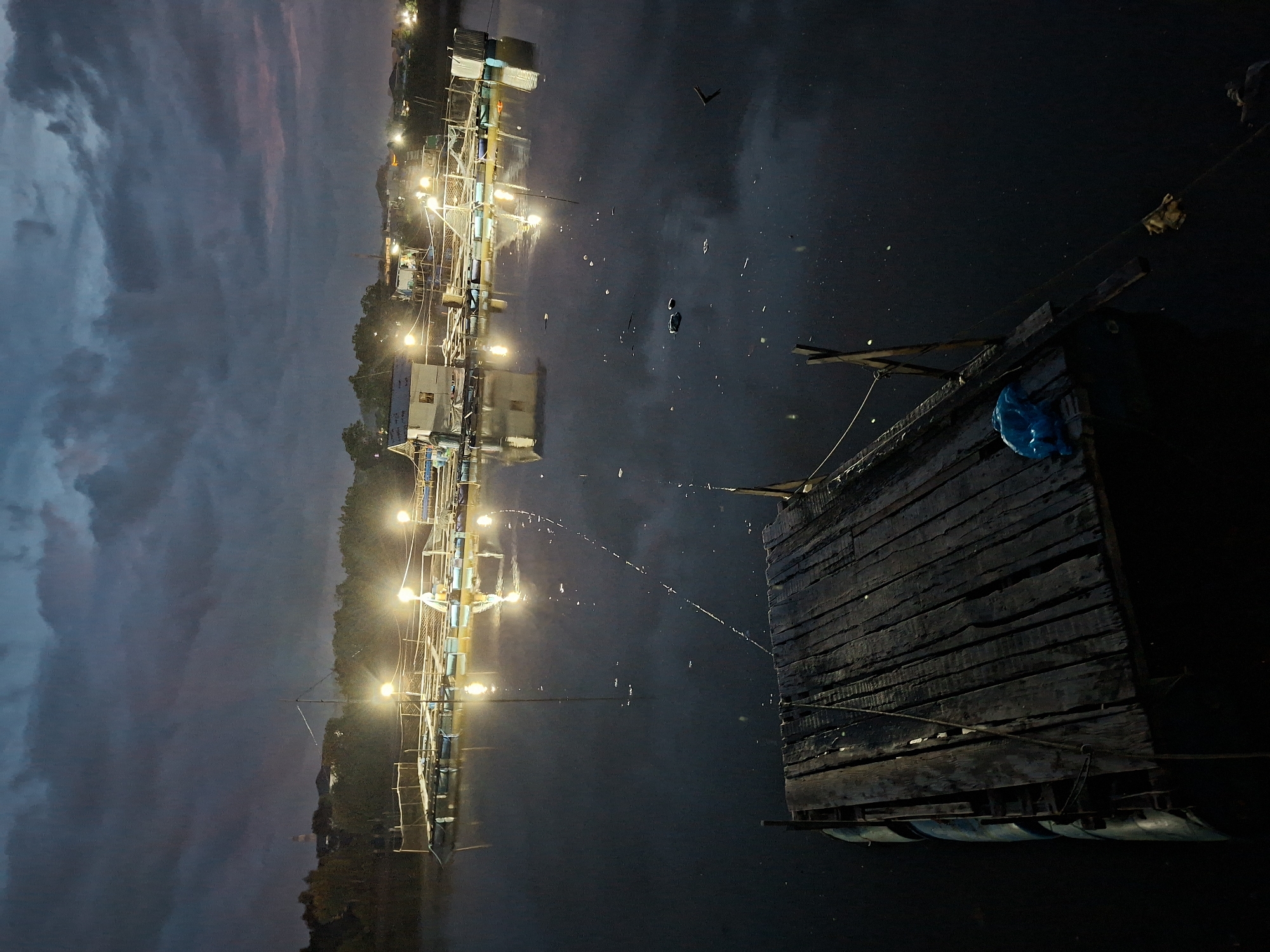 The image captures a night scene in Lhokseumawe, Indonesia. A wooden raft floats on water, with a blue plastic bag visible on its structure. A network of illuminated structures, possibly buildings or platforms, is reflected in the water, suggesting a calm, still surface. The background sky is a mix of dark clouds with hints of dusk or dawn colors.