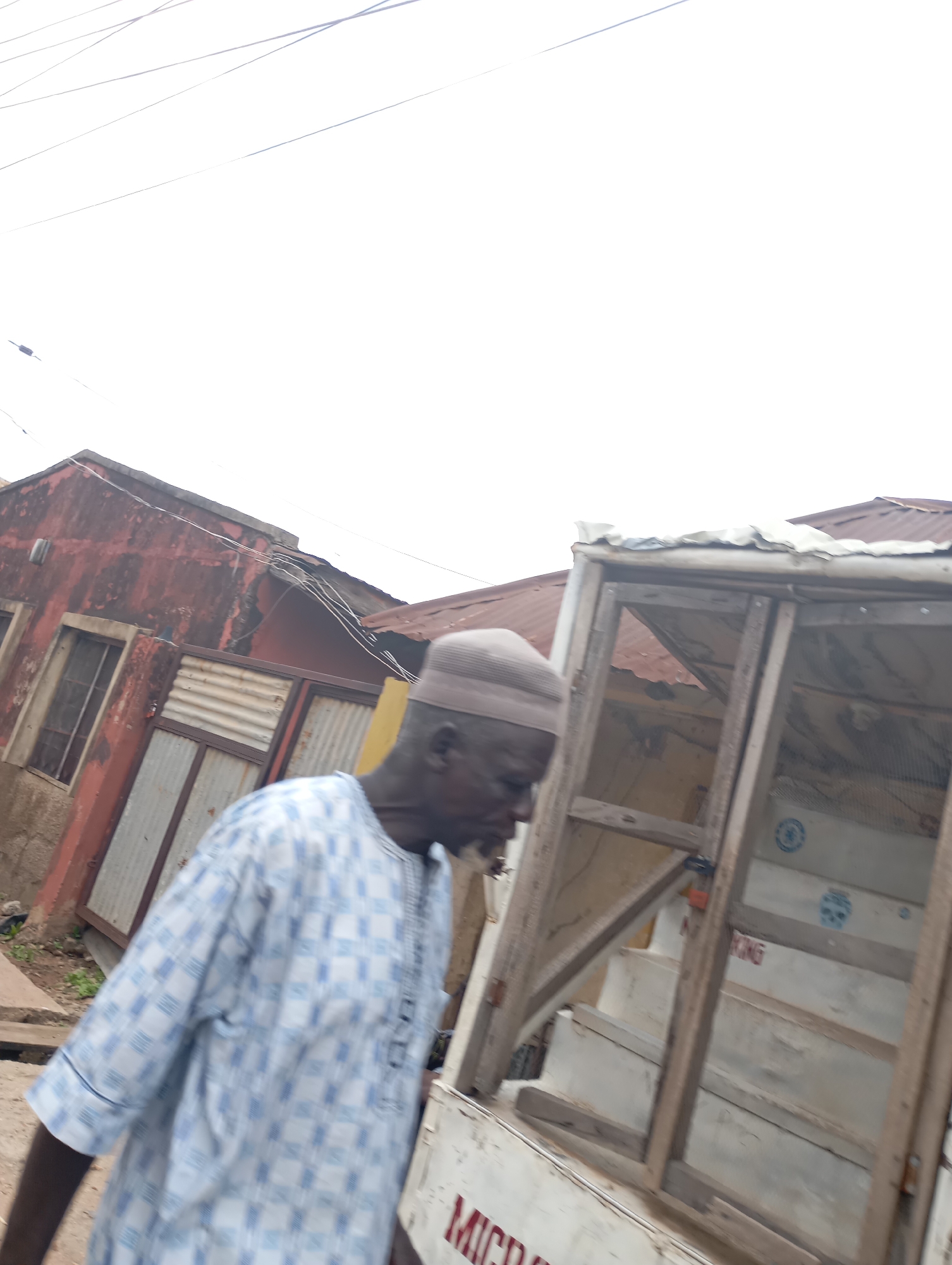 An outdoor, medium shot captures an elderly man walking past a wooden structure with mesh screens in Shendam, Nigeria. The man, wearing a light brown cap and a light blue and white checkered traditional tunic, is positioned on the left side of the frame. He is looking towards the right. The wooden structure, which appears to be a mobile cart or stall, occupies the right and central portions of the image. Its frame is constructed from light-colored wood, and it features open sections covered with mesh. The background reveals a weathered, red building with corrugated metal siding and windows, suggesting a residential or commercial area. Overhead, electrical wires are visible against an overcast sky. The ground appears to be unpaved. Part of the word "MICR" is visible on the side of the cart.