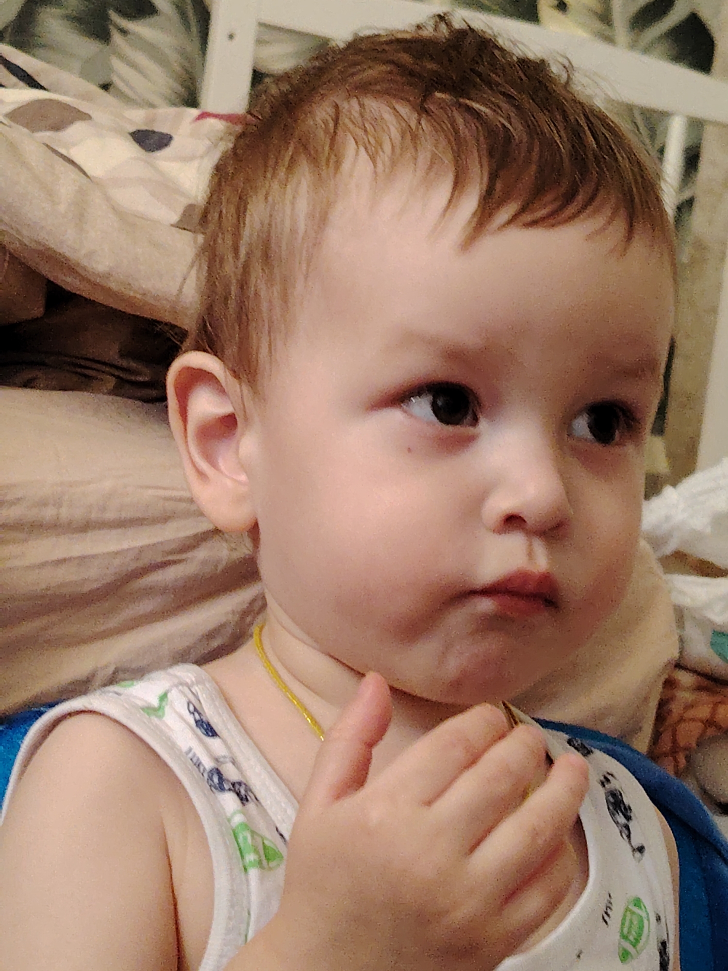 The image displays a close-up of a young child in an indoor setting within the city of Марковский, Russia. The child, possessing light skin, brown hair, and dark eyes, is positioned with their head turned slightly left, and their gaze directed upwards and to the right. A white sleeveless undershirt, patterned with blue and green motifs, is worn. A thin, gold-colored chain is visible around the child's neck. The child's left hand, with fingers partially curled, is positioned near the chest. The mouth exhibits a slight downturn. The background consists of soft textiles; specifically, a light-colored pillow featuring irregular brown and dark blue patterns is on the left, and a textured light brown fabric is on the right. A white structural element is partially visible in the upper background.