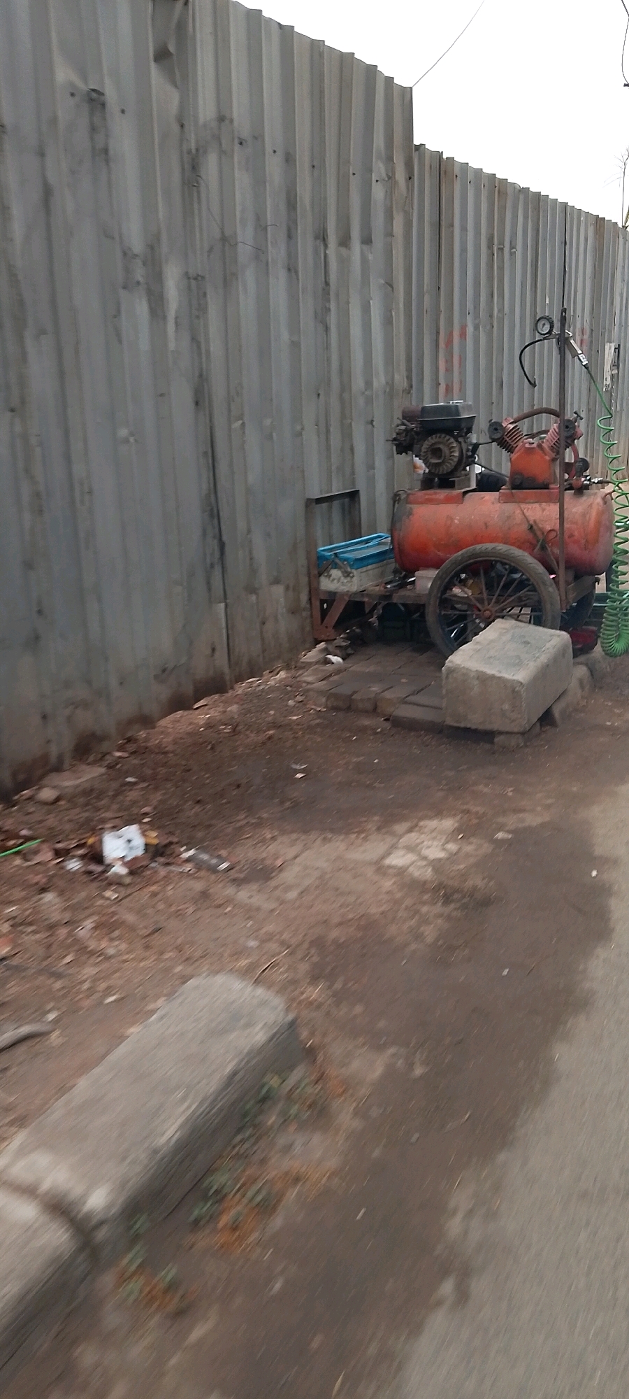 In an outdoor setting, an orange air compressor with a black engine sits next to a corrugated metal fence in what appears to be Bekasi, Indonesia. The air compressor is mounted on a wheeled frame and has a pressure gauge. A green coiled hose is attached to it. Beside the air compressor is a blue plastic container on a stand made of metal and wood. The ground is a mixture of dirt and paving stones, with some debris scattered around. In the foreground, a concrete curb is visible, and a paved surface extends into the background. The lighting suggests it is daytime.