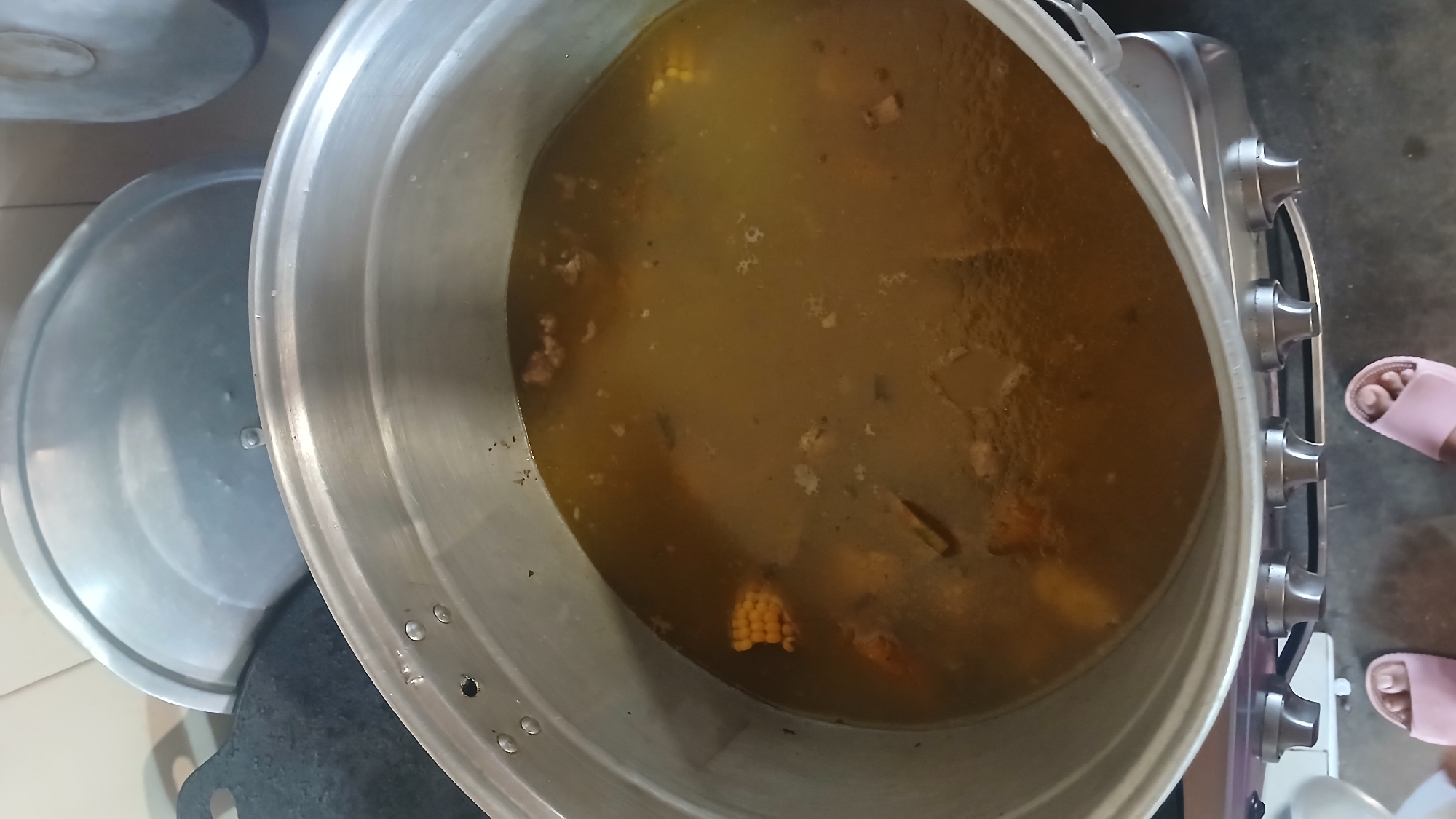 The image shows a large, open aluminum pot filled with a simmering, brownish liquid, likely soup or stew, containing visible ingredients such as corn and pieces of meat or vegetables. The pot rests on a stovetop with silver knobs. To the left of the pot, there is a separate pot with its lid ajar, both also appearing to be aluminum.
The scene appears to be indoors, likely in a kitchen. Parts of a person's feet, wearing pink flip-flops, are visible in the lower right corner of the image.
Given the prompt's specification, this scene is said to be in the city of Altagracia, Venezuela.