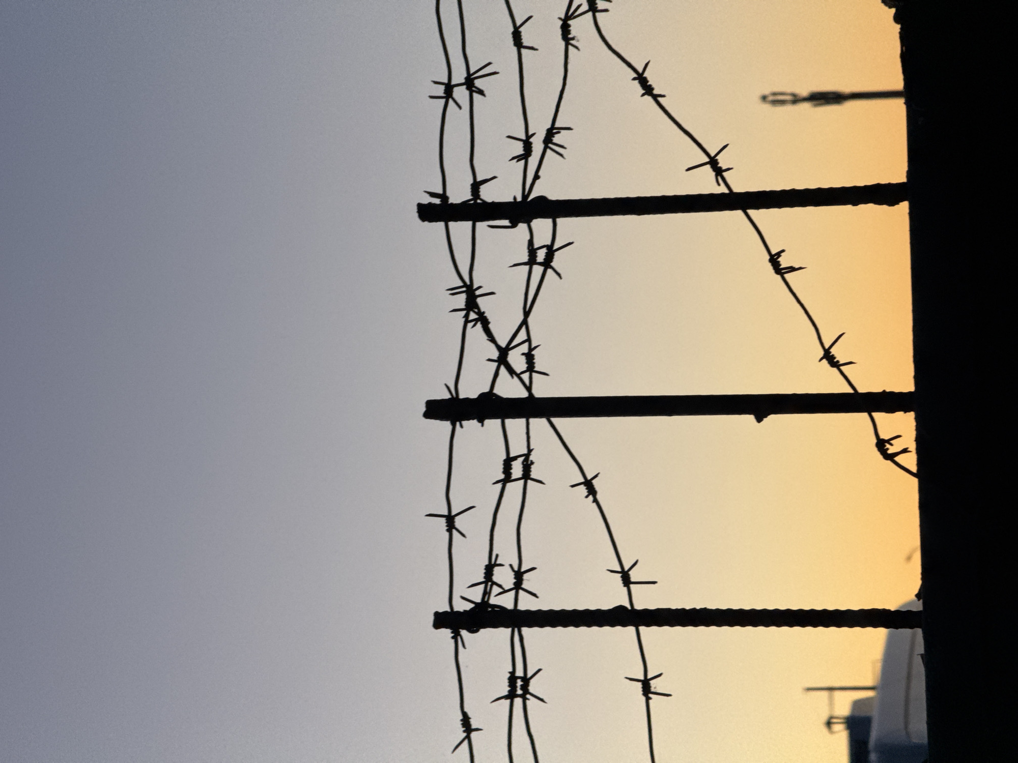 The image depicts a barbed wire fence against a sunset or sunrise sky. The fence consists of metal posts with barbed wire strands stretched between them. The wire and posts are silhouetted against the sky, which transitions from a lighter color on the left to a warmer orange on the right. Some buildings are visible through the fence in the background. The location, Minsk, Belarus, is not evident in the image itself and is not verifiable.