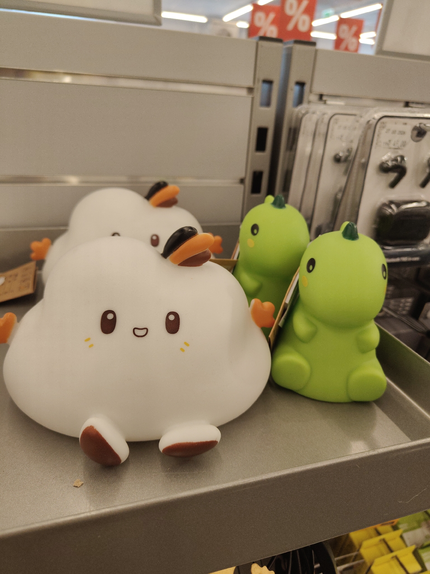 This is a close-up, eye-level shot of a retail display in a store, possibly a discount store, in Nowy Dwór Mazowiecki, Poland. The focus is on several novelty lamps in the foreground. On the left are two white, cloud-shaped lamps with simple cartoon faces, each with two brown eyes and a smiling mouth, adorned with orange and black duck-like accessories. To the right are two bright green dinosaur-shaped lamps, also with cartoon eyes. The items are arranged on a gray shelf. In the background, out of focus, are rows of merchandise in clear plastic packaging and red sale signs with "%" symbols. The lighting is bright and slightly diffused, typical of a retail environment.
