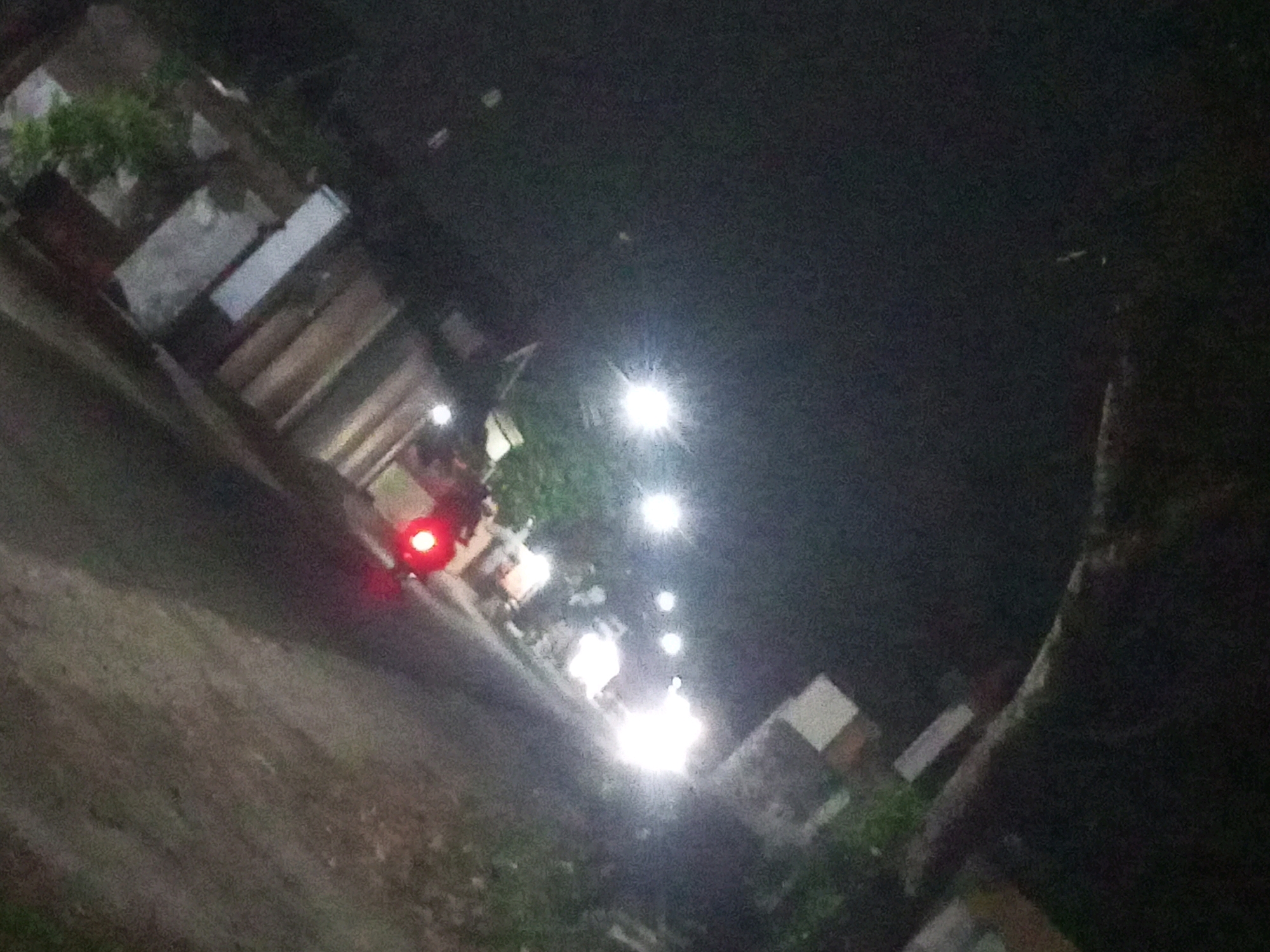 The image shows a dimly lit street at night, possibly in the city of Ngasem, Indonesia. Several illuminated light sources are visible, including streetlights, indicating an urban or semi-urban setting. In the foreground, a motorcycle with its red taillight illuminated is positioned on the left side of the frame. Structures that appear to be buildings or stalls line the right side of the road, some with visible lights. The ground is a rough, unpaved surface. The overall scene suggests a quiet night on a local street.