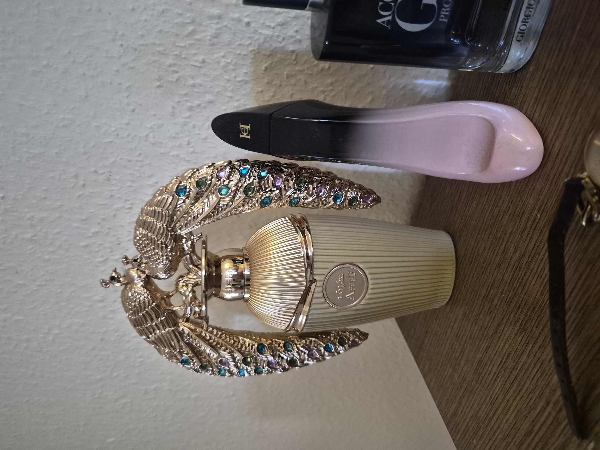 This media file displays a collection of perfumes on a wooden surface, with a textured white wall in the background. The main subject is an ornate perfume bottle with a gold, peacock-feather-like design encrusted with colorful gems. The bottle itself is a pale gold or cream color with vertical ribbed detailing on its body and a silver cap. The peacock design appears to be made of metal and is intricately detailed, with small bird heads at the top of the feathers.
To the right of the peacock perfume bottle, another perfume bottle is visible. This bottle is black at the top and fades into a pale pink color, with a sleek, curved design and a gold logo on its side that appears to be a stylized "CH".
Further in the background, a dark-colored perfume bottle is partially visible, with some text that reads "ACQUA" and "GIORGIO".
The lighting suggests it may be daytime, but there are no clear indicators of the time of day or weather. The setting appears to be indoors, likely on a shelf or vanity. No people are visible in the image, and the focus is solely on the perfume bottles. The overall impression is one of luxury and decorative display.