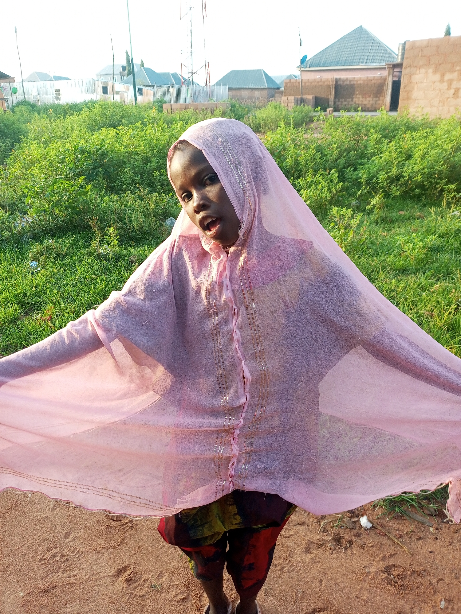 A young Black girl wearing a pink hijab and a colorful skirt stands outdoors in Tempure, Nigeria. She is positioned in the center of the frame, facing the camera with her mouth open. Her hijab is sheer and adorned with sparkly embellishments. The girl is surrounded by lush green vegetation and stands on a sandy, reddish-brown surface. In the background, there are buildings with corrugated metal roofs and brick structures, indicative of a residential area. A radio tower is visible in the distance. The lighting suggests it is daytime, possibly late afternoon or early morning, with the sun casting a warm glow.