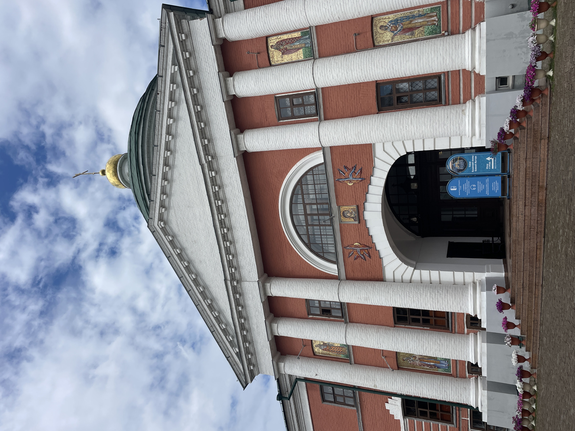 The image shows the exterior of the Kazan Mother of God Monastery in Kazan, Russia. The building is made of red brick and features white columns and a white pediment. A golden dome with a cross tops the structure. Mosaic religious icons adorn the walls between the columns. An arched entryway leads into the building, with steps leading up to it. Potted flowers line the steps and the walkway in front of the building. Two blue signs stand on the steps, providing information about the monastery. The sky above is partly cloudy.