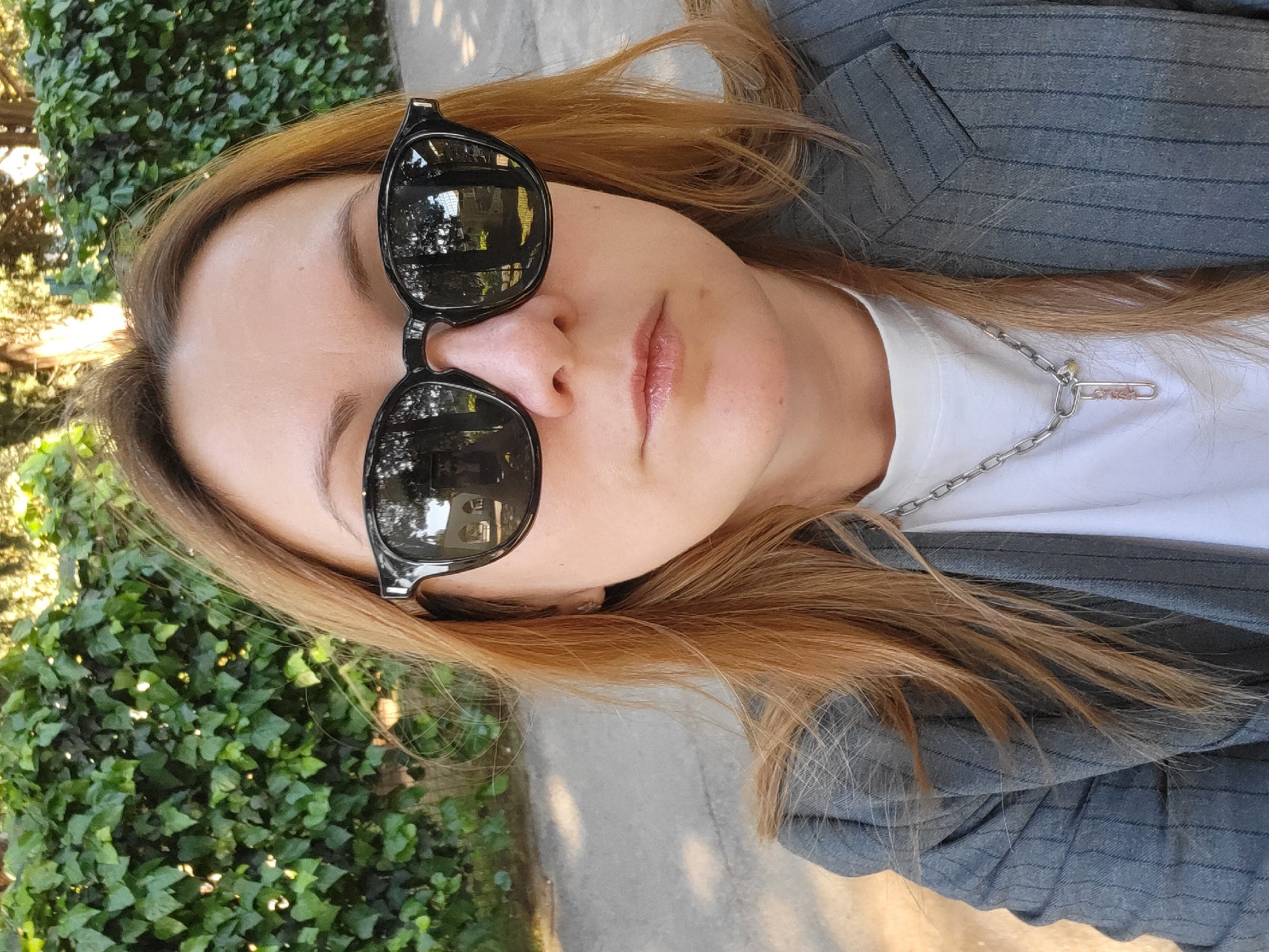 This close-up, slightly angled portrait features a person, likely female, with long, light brown hair styled over her shoulders. She wears black sunglasses that reflect trees and what appear to be distant buildings. Her lips are slightly parted with a glossy finish. She is dressed in a white t-shirt, over which a silver chain necklace with a distinctive, elongated, paperclip-shaped pendant is visible. A grey pinstriped blazer or jacket completes her attire.
The setting is outdoors in Budva, Montenegro, with dense green foliage, possibly ivy, visible on the left side of the frame. A light-colored paved path or sidewalk runs along the right side and behind her. The bright, natural light and dappled sunlit patches on the foliage indicate it is daytime and sunny. The person appears to be posing for a photograph, likely a selfie, in a relaxed manner. No clearly legible text is visible on the clothing or accessories.