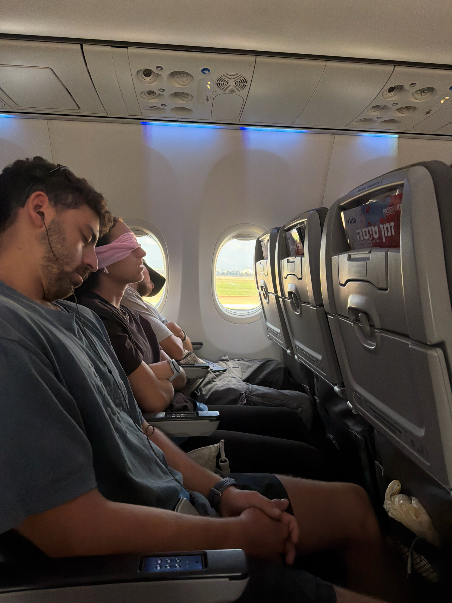 The image depicts the interior of an airplane cabin, likely on a commercial flight. In the foreground, a man with a beard and headphones is asleep, wearing a gray t-shirt. To his right, a person is wearing a pink blindfold and a dark shirt, also appearing to be asleep. Behind them, another person in a light-colored shirt and a black baseball cap is visible, also asleep. The seats in front of them show tray tables and in-flight entertainment screens. Above the seats, air vents and overhead compartments are visible, with blue LED lighting illuminating the ceiling. Outside the window, a landscape of green fields and a bright sky suggests a daytime flight. The presence of Hebrew text on a magazine visible in the seat pocket of the second row ("Tman Simona") indicates that this flight is likely operating to or from Israel. Yehud Monosson is a city in Israel, and this scene could represent passengers on a flight arriving at or departing from Ben Gurion Airport, which is located near Yehud Monosson.