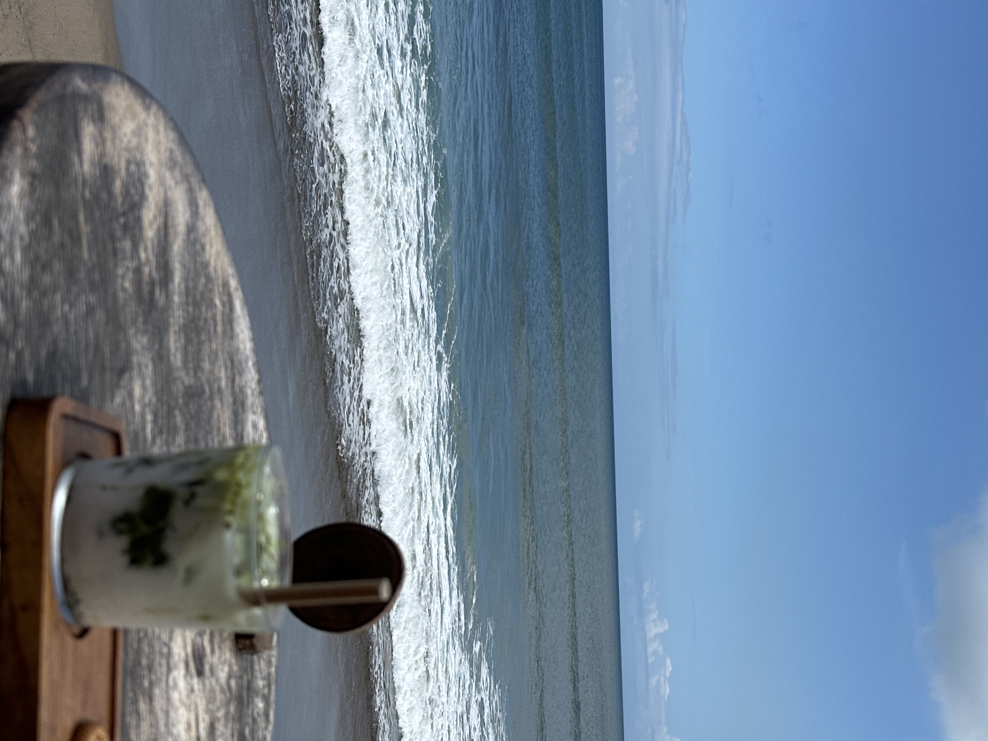 The image shows a glass of a matcha drink on a wooden table with a view of the beach. The location is in North Kuta, Indonesia, as the beach and clear blue sky are consistent with the area. The waves are visible as they come to the shore, and the sky is blue with a few clouds.