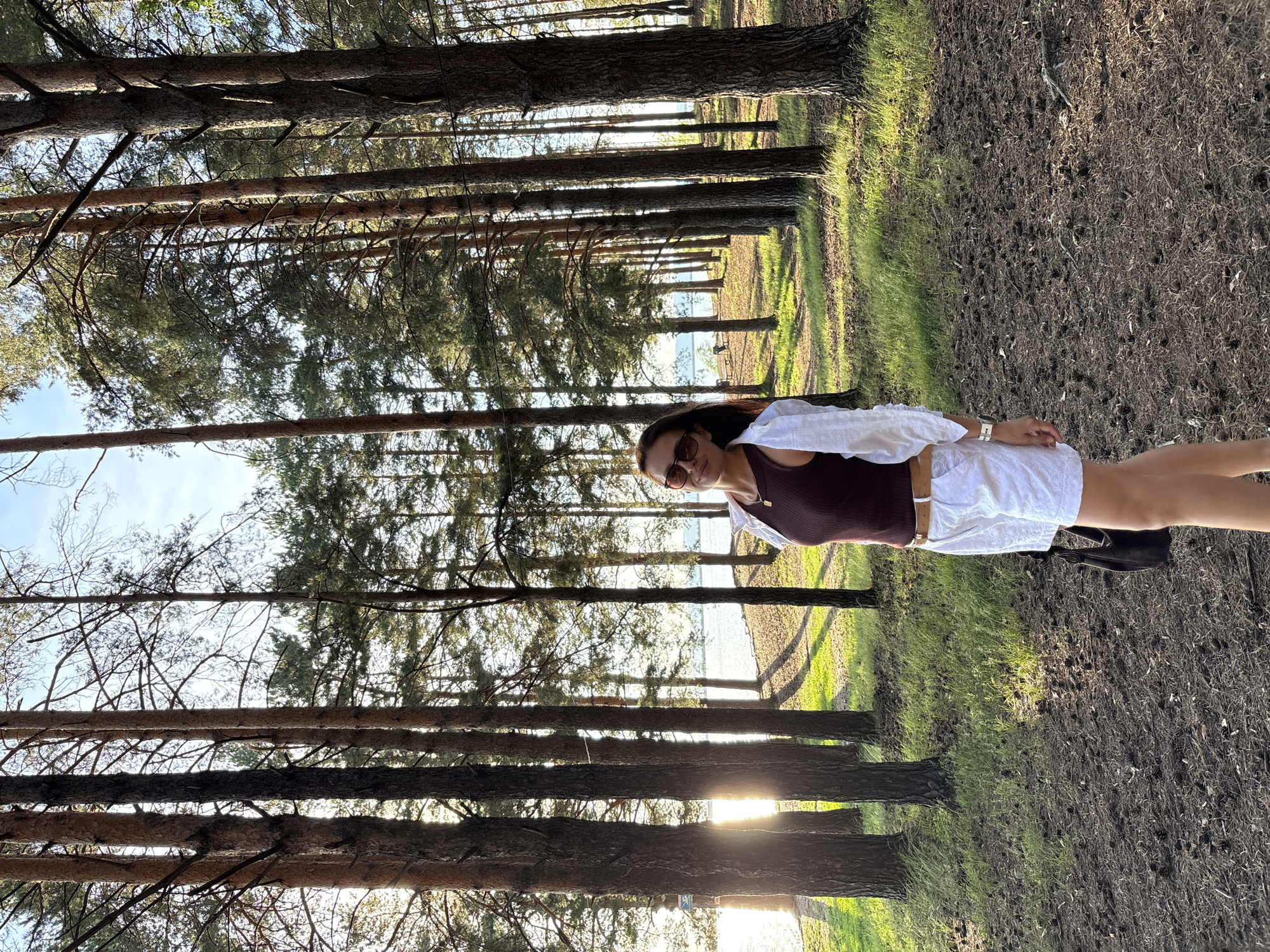 A woman is posing for a picture in a pine forest. She is wearing a burgundy tank top, a white open-front shirt, and white shorts. She has on sunglasses and a watch. The woman is standing with her right side facing the camera, and her head is turned towards the camera as she smiles. The sun is shining through the trees, creating lens flare. The ground is covered in pine needles and some green grass. In the background, there are more pine trees, and a body of water is visible, possibly a lake or river. The scene appears to be set in the late afternoon or early evening, given the warm, golden light. The location is Chaikovsky, Russia. There is no visible text in the image.