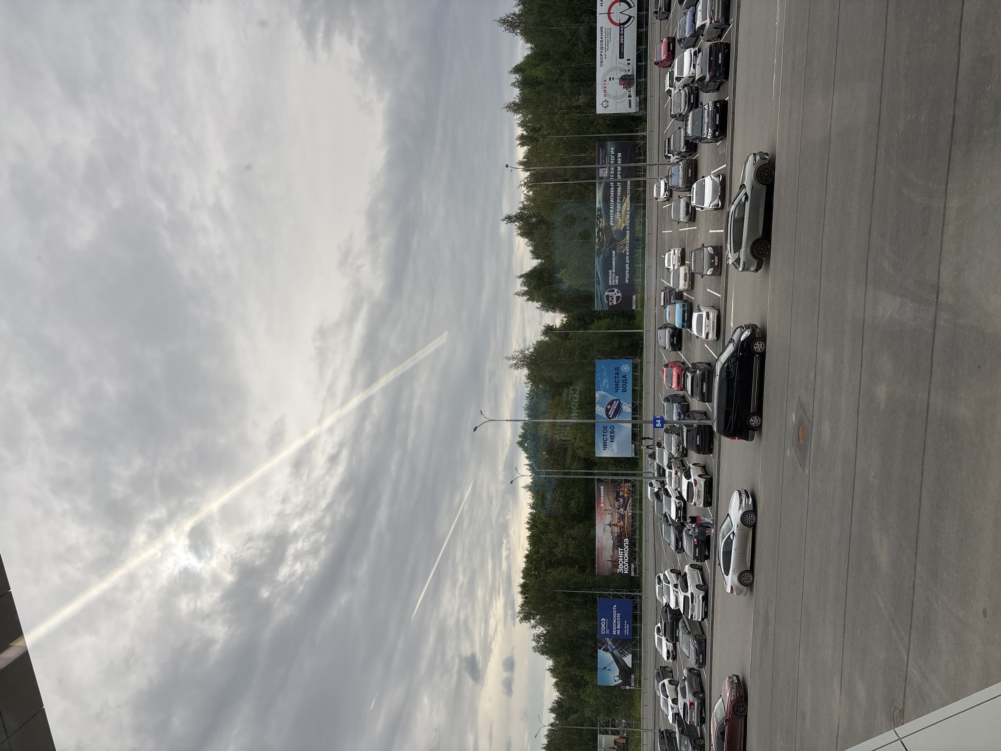 This image shows a parking lot filled with cars, viewed from an elevated angle. The cars are parked in rows, suggesting a public space like a shopping center or event venue. In the background, there is a line of trees and a cloudy sky with a distinct contrail from an aircraft. Several billboards are visible along the edge of the parking lot, featuring Russian text related to various businesses and services, including "Союз" (Union), "Безопасность на высоте" (Safety at height), "Чистое небо" (Clear sky), and "Чистая вода" (Clean water). The overall scene appears to be a daytime setting, with the lighting suggesting it might be overcast or late afternoon. The presence of multiple cars and billboards indicates a commercial or public area.