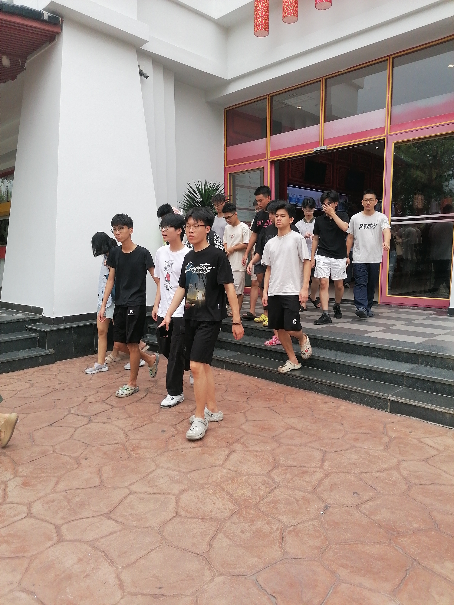 A group of young people are walking down the steps of a building. The building has a modern facade with large glass windows trimmed with red and gold. Red lanterns hang above the entrance. The steps are dark grey with a textured, reddish-brown concrete walkway in front. The setting appears to be an outdoor public space in Bai Chay Ward, Vietnam. The group consists of about nine people, dressed in casual clothes such as t-shirts, shorts, and sandals. They are walking together, suggesting they may be a group of friends or acquaintances.