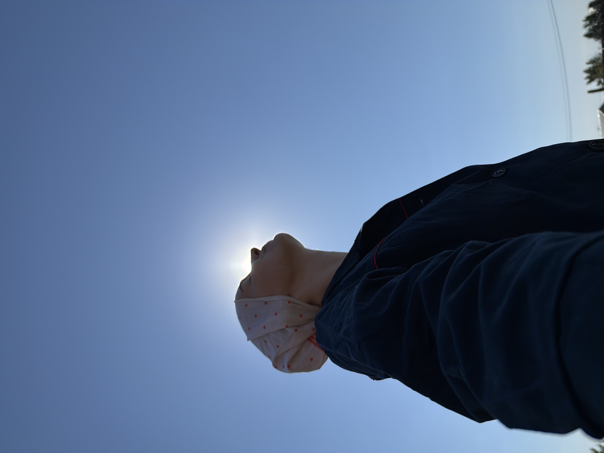 A person wearing a polka dot bandana and a dark jacket is looking directly at the sun in a clear, blue sky. The photograph appears to be taken from a low angle, capturing the silhouette of the person against the bright sunlight. The location, Chaikovsky, Russia, is not evident within the image. The background is primarily the solid blue of the sky with some vegetation at the top right.