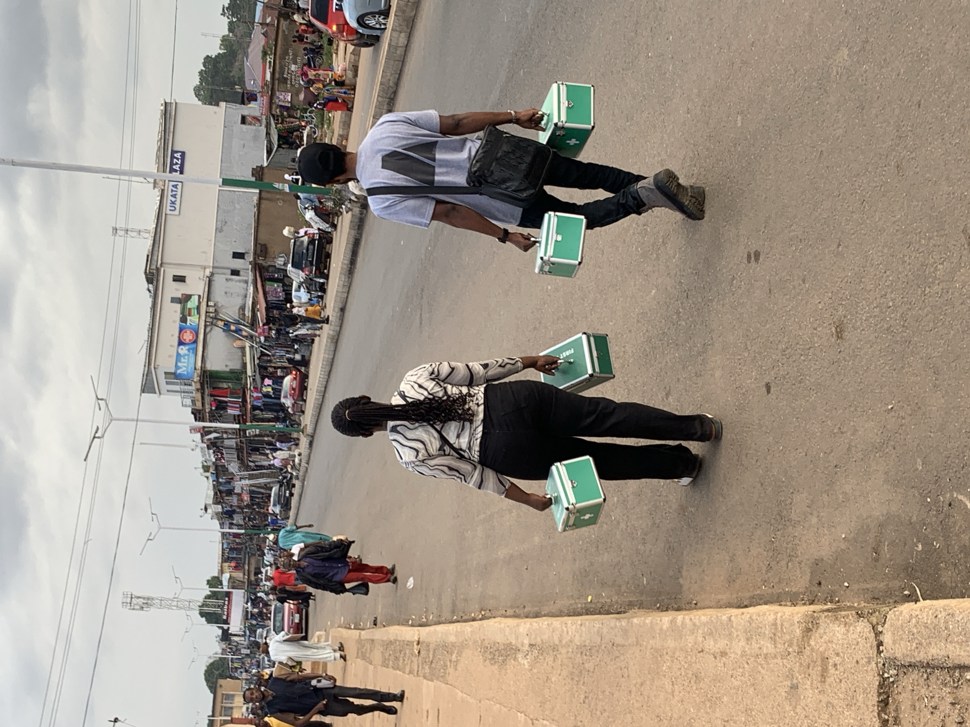 Two individuals are walking on a paved road in a busy market area in Jos, Nigeria. The person in the foreground, on the right, is a woman wearing a black and white striped long-sleeved shirt and black pants. She is carrying two small, green medical kits with white crosses on them. The person in the background, on the left, is a man wearing a grey t-shirt, black pants, and a black cap. He is also carrying two similar green medical kits and has a black messenger bag slung across his body.
The scene appears to be a street market with shops and stalls lining the road, indicated by the buildings and merchandise visible in the background. There are other people walking and some vehicles present, suggesting a bustling daytime environment. The sky is overcast, indicating cloudy weather. The overall atmosphere seems to be that of daily commerce and pedestrian activity. The "UKATA PLAZA" text is visible on a building in the background. The medical kits suggest that these individuals may be involved in healthcare services or carrying supplies for such.