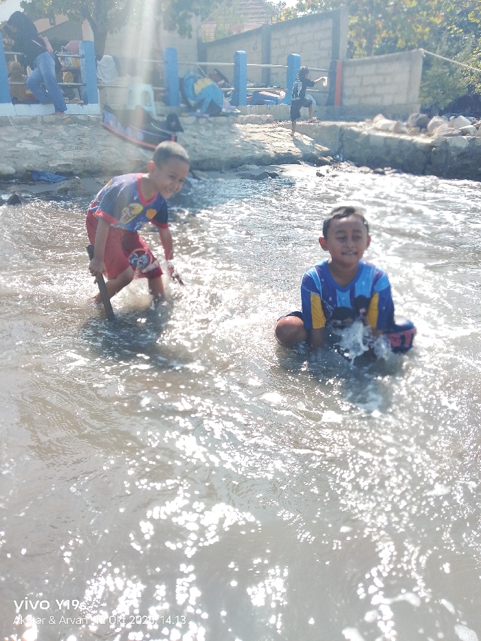 This image depicts a daytime scene in Limbangan, Indonesia, at a body of water bordered by a concrete bank. In the immediate foreground, two young boys are playing in shallow, rippling water under bright sunlight, which creates significant reflections and lens flare. The boy on the left wears a blue and red t-shirt with red shorts, standing and actively splashing water while holding a stick. He is looking towards the viewer and smiling. The boy on the right, also smiling towards the viewer, is seated or kneeling in the water, wearing a blue and yellow long-sleeved shirt and dark shorts, with water splashing around his chest area.
The midground features a concrete bank or platform elevated above the water level. Blue-painted poles form a railing along this bank. A small, black and red toy boat is visible on the concrete surface. Several individuals are present on this bank: one person in a black top and blue jeans stands on the left, another person is seated to their right, and a child in a dark shirt stands near a light-colored concrete block wall further right. Assorted items, possibly clothing or bags, are scattered on the bank. The background reveals a concrete block wall and sparse vegetation under a bright sky.