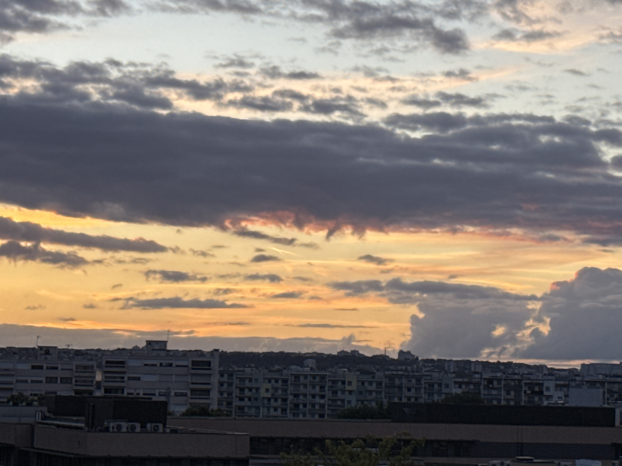 The image depicts a city skyline at dusk, as indicated by the dramatic cloud formations painted with sunset hues of orange, pink, and purple. The sky transitions from a pale blue at the top to a vibrant, warm palette closer to the horizon. Dark, heavy clouds dominate the upper and middle portions of the sky, creating a stark contrast with the lighter, illuminated areas. Below the sky, a densely populated urban landscape unfolds. Numerous multi-story buildings, appearing to be residential or office blocks, are visible, characterized by rows of windows and a uniform, somewhat monolithic architectural style. The buildings are densely packed, suggesting a significant urban area. In the foreground, closer structures with flat roofs and visible air conditioning units indicate a layered cityscape. The overall impression is of a large, modern European city during twilight, with an emphasis on the atmospheric conditions of the sky and the architectural density. This scene is consistent with views from the city of Fresnes, France, known for its residential developments and suburban character.