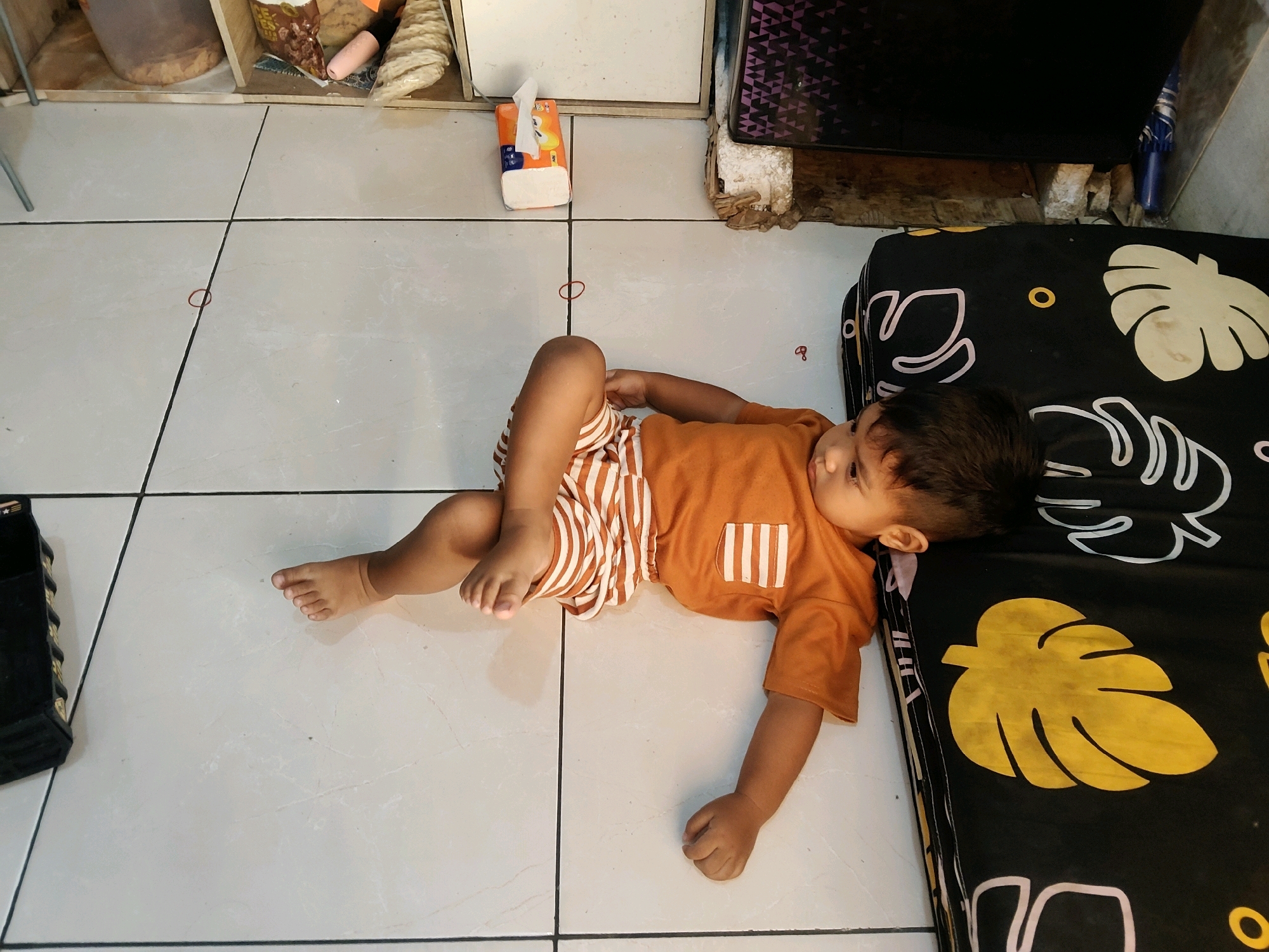 A young child is lying on the floor next to a black mattress with yellow leaf patterns. The child is wearing an orange t-shirt with white vertical stripes on the pocket and striped orange and white shorts. The child is lying on their left side with their head resting on the mattress and their eyes open. The floor is tiled with white tiles, and there are dark grout lines. In the background, there is a tissue box and some shelves with various items. The scene appears to be indoors, possibly in a residential setting within Mekarmukti, Indonesia.