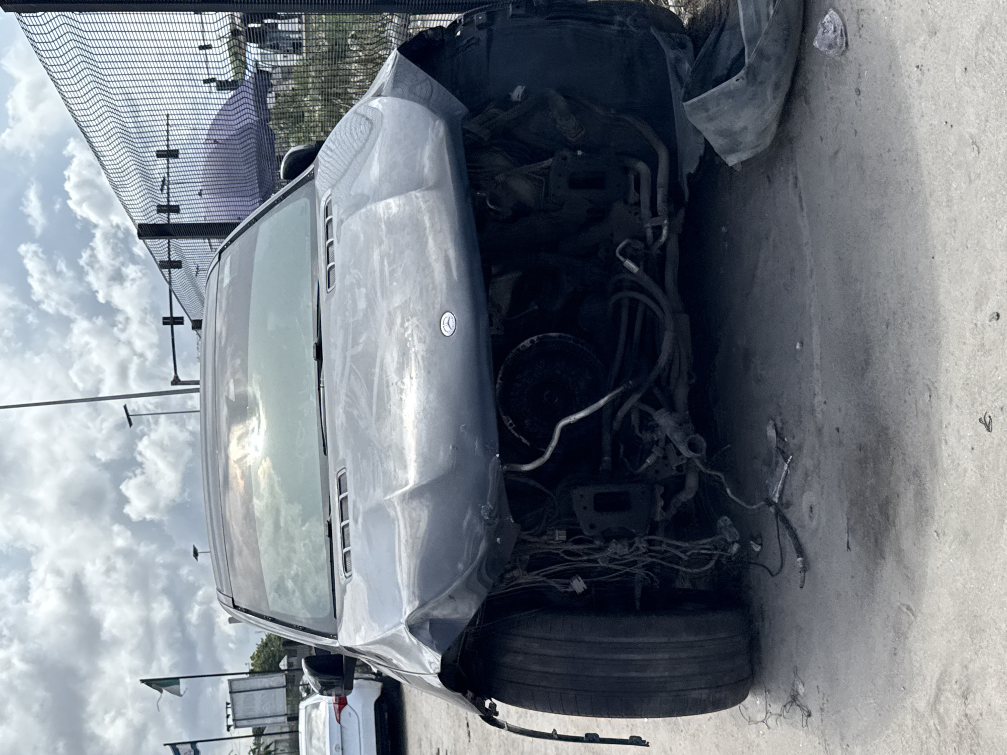 The media file displays a severely damaged gray Mercedes-Benz SUV, viewed from its front-left angle, resting on what appears to be a concrete or asphalt surface. The vehicle has sustained extensive front-end damage, with the bumper and grille missing or completely destroyed, leaving the engine bay exposed. Wires, hoses, and various engine components are visible within the damaged front section. The Mercedes-Benz emblem is still distinguishable on the crumpled hood.
The scene is outdoors during the daytime, under a cloudy sky. A tall, dark metal fence or netting structure runs along the top of the image, suggesting an enclosed area like a junkyard, salvage yard, or vehicle storage facility. In the background, other partially visible vehicles, including a white one, and some poles or structures can be discerned. A small white flag is also visible in the distance. The ground immediately around the car is somewhat uneven and dusty. There are no people visible in the image, and no specific activity beyond the static display of the damaged vehicle is depicted. The overall context suggests the car has either been involved in a major accident or is undergoing dismantling for parts in Igboefon, Nigeria. No significant text is visible apart from the car's branding.