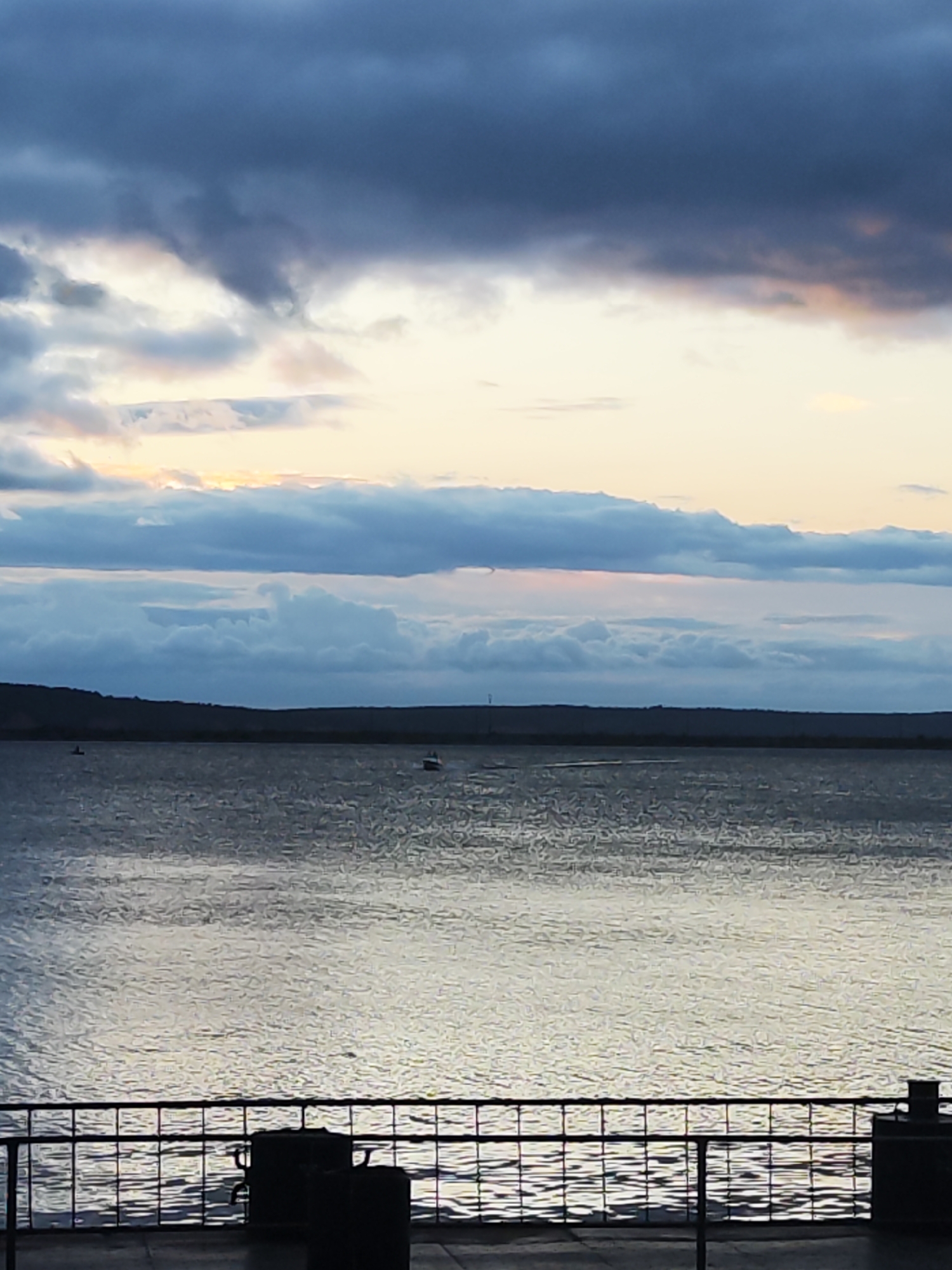 The image captures a serene waterscape in Chaikovsky, Russia, likely during twilight given the soft, diffused light. A vast body of water, possibly the Kama River or a reservoir, dominates the scene, its surface rippled and reflecting the muted colors of the sky. In the distance, a dark, undulating shoreline separates the water from the sky. The sky itself is a dramatic canvas of clouds, with darker, heavy formations at the top transitioning to lighter, wispy layers tinged with the warm hues of sunset near the horizon. A few small boats are visible on the water, hinting at recreational activity. In the foreground, a dark metal railing creates a barrier, suggesting a viewpoint from a promenade, pier, or a similar elevated platform. Silhouetted against the water are some indistinct, dark objects, possibly urban furniture or structures, adding a touch of human presence to the natural landscape. The overall impression is one of peaceful contemplation, with nature's grandeur on full display.