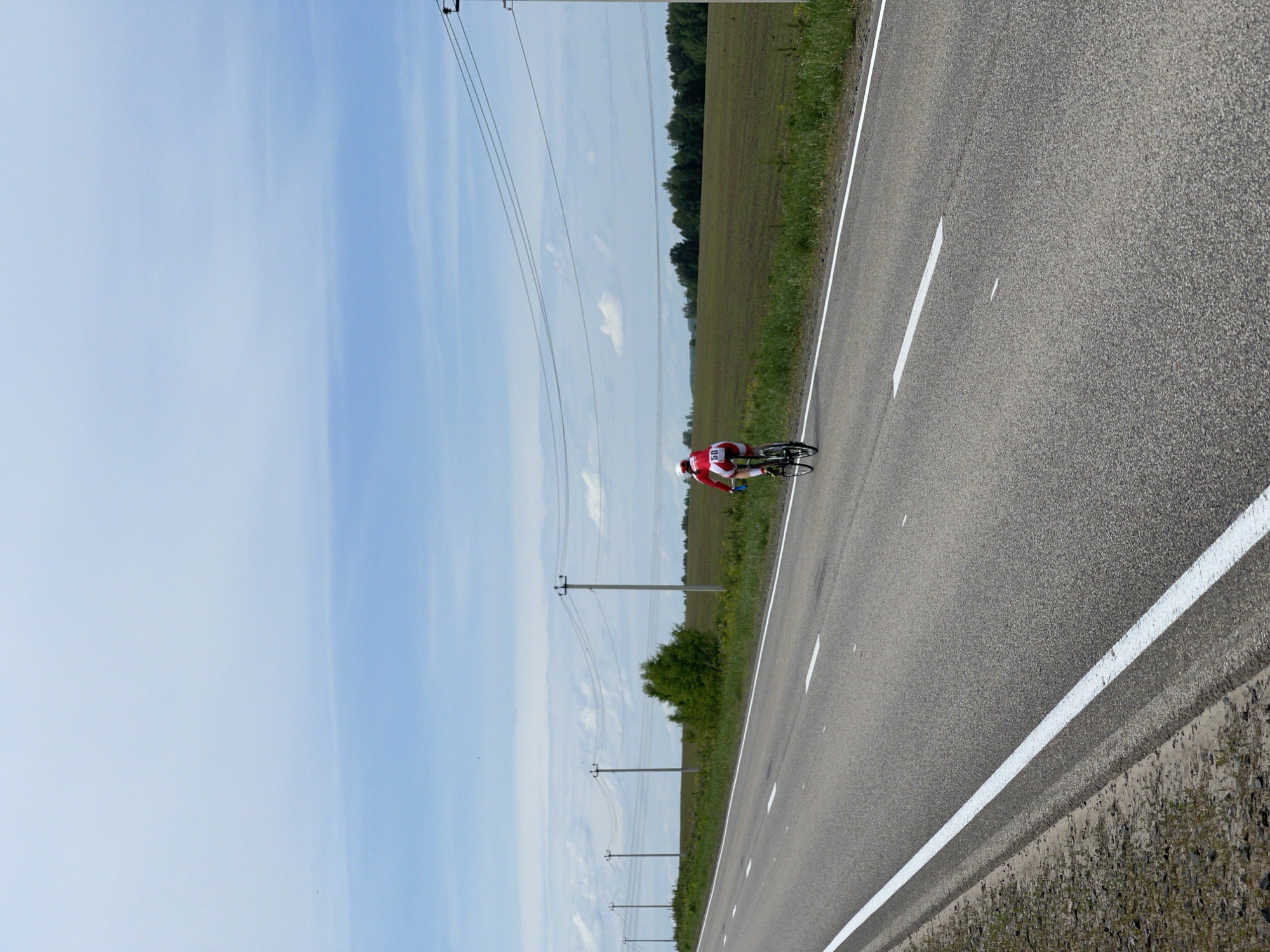 A cyclist in a red and white outfit rides on an asphalt road marked with white lines. The sky is pale blue with wisps of white clouds. Power lines and poles run parallel to the road along a field of green vegetation. The image is taken in Maksimovo, Russia.