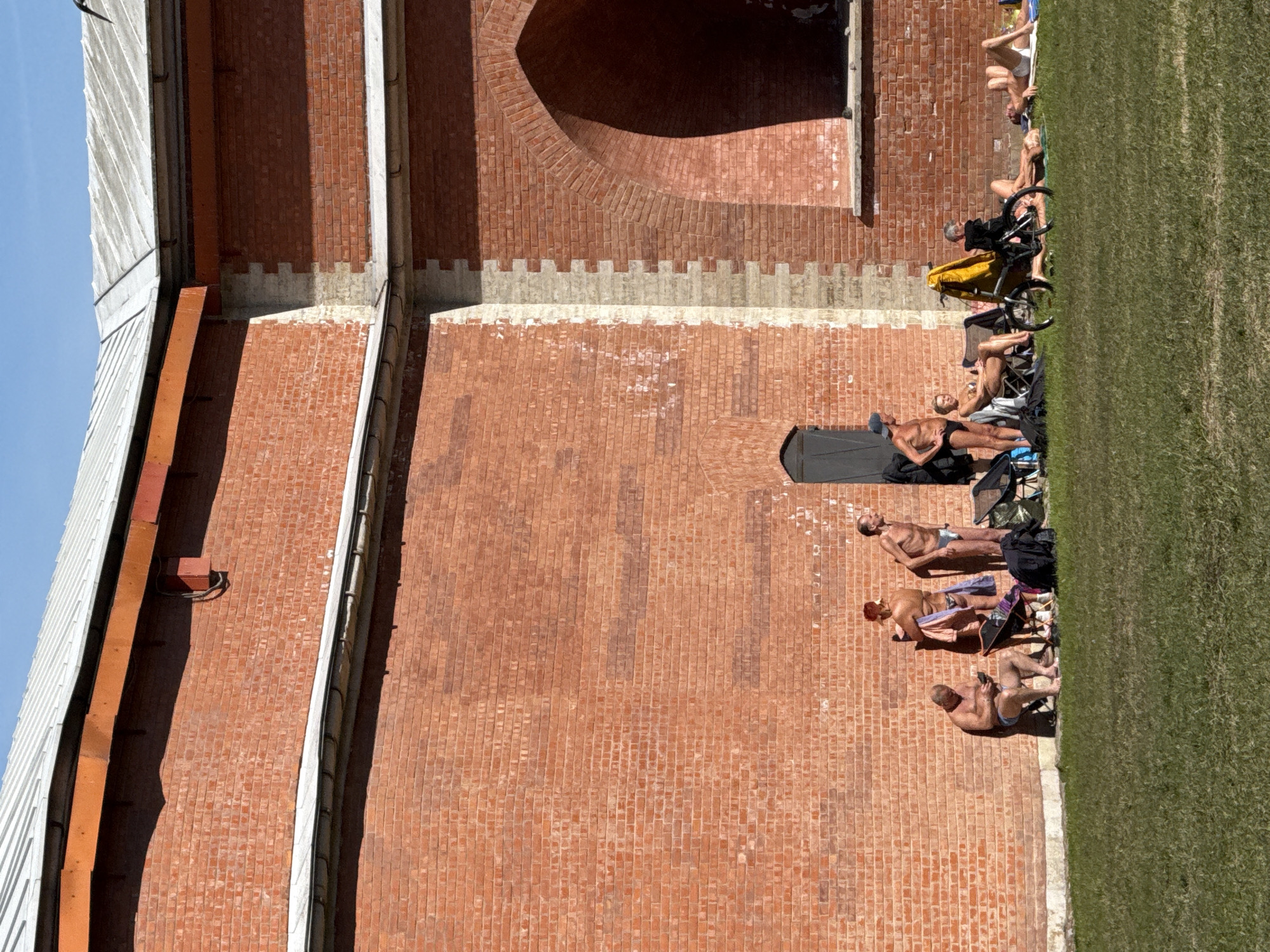 The image depicts a group of individuals sunbathing on a strip of grass adjacent to the brick wall of the Peter and Paul Fortress in Saint Petersburg, Russia. The fortress wall is constructed of red bricks with a large archway visible near the top. Individuals are lying on towels and chairs, some shirtless, near the edge of a green lawn, and some have bicycles nearby.