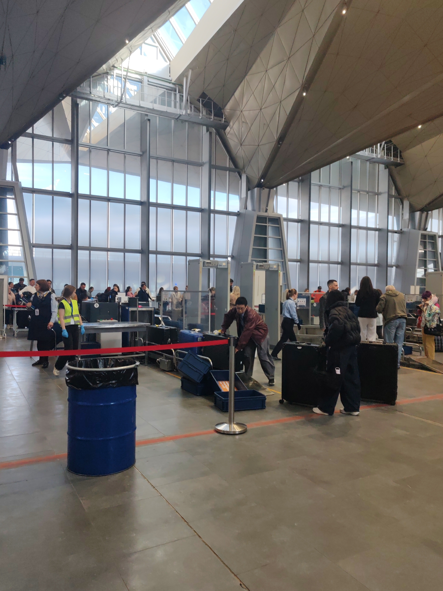 The image captures an active scene inside a modern airport terminal, identified as being in Saint Petersburg, Russia. The setting features a high, geometrically patterned ceiling with large, angular light-colored panels, supported by substantial pillars. Extensive glass walls line the perimeter, allowing abundant natural daylight to illuminate the space, suggesting it is daytime with clear skies outside. The floor is covered with large grey tiles.
The main activity centers around an airport security checkpoint or baggage drop-off area. Numerous people, both passengers and airport staff, are visible. Several grey metal detector arches are positioned in the background, with passengers queuing to pass through them. In the foreground and midground, passengers are handling their luggage, preparing it for screening or check-in. One individual in a dark jacket is seen bending over a large black suitcase. Airport staff members, some in dark uniforms with light blue shirts and others in bright yellow high-visibility vests and blue gloves, are present, guiding passengers and overseeing the process.
Prominent objects include various suitcases, primarily in dark colors like black and blue, scattered on the floor and on tables. A bright blue metal barrel with a black trash bag liner serves as a trash can in the left foreground, next to a red fabric barrier tape that delineates a path or boundary. Silver stanchions also contribute to crowd control. In the background, a black screen or monitor shows a white number "9." The scene conveys a typical, moderately busy airport environment with people moving purposefully through the required procedures.