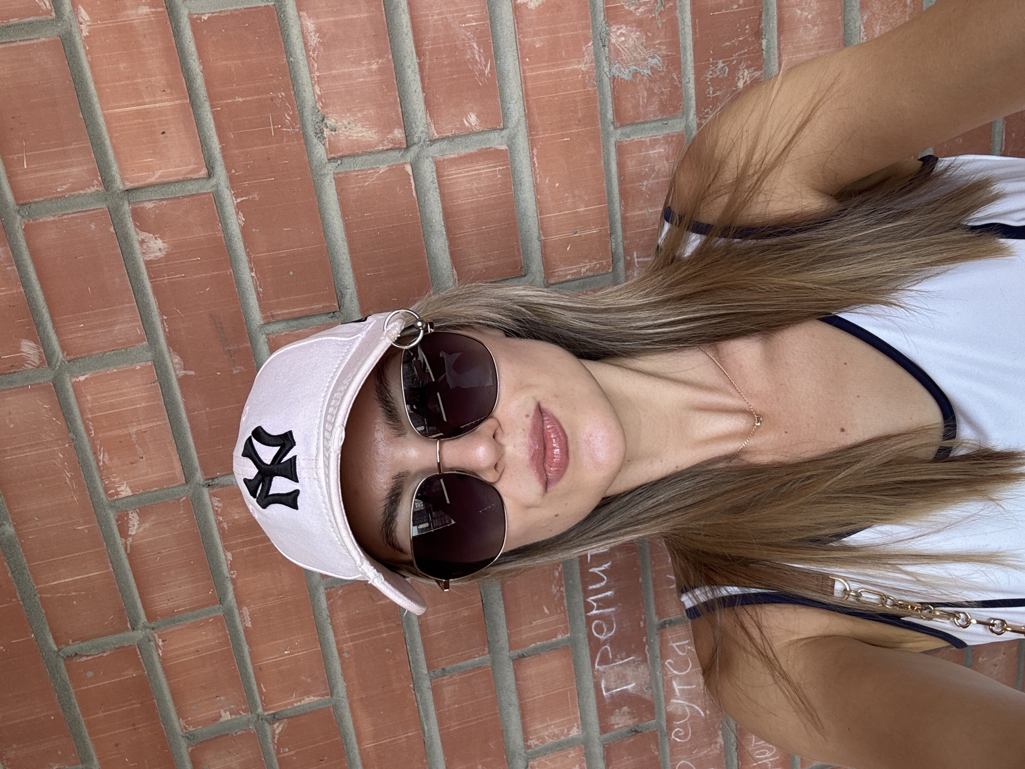 A woman in a white and navy blue tank top and a white baseball cap with a black logo is wearing sunglasses. The background is a brick wall, and Cyrillic writing is on the bottom of the wall. The city and specific location are unknown.