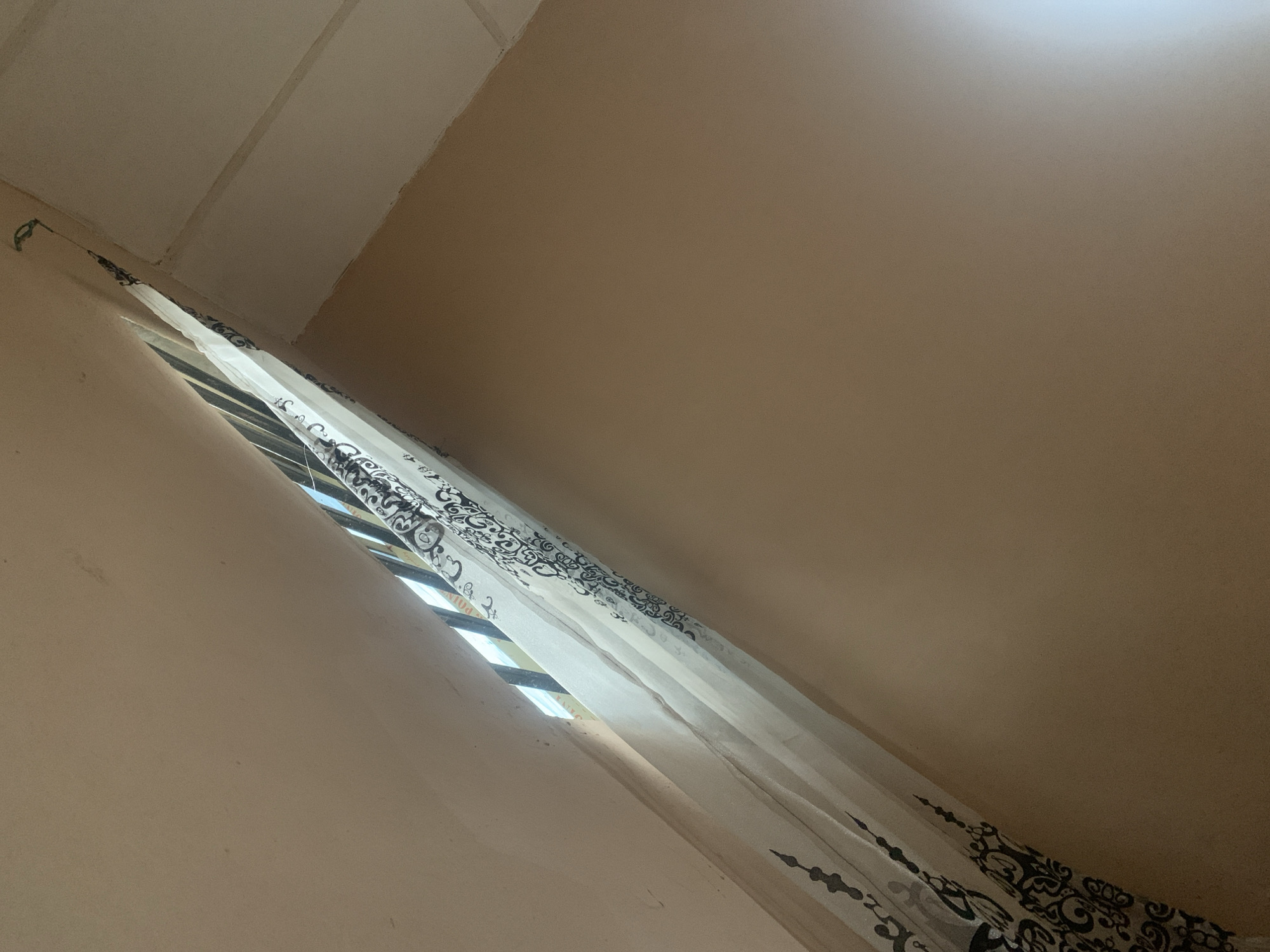 An interior shot looks up towards a corner where two walls and a ceiling meet. A sheer white curtain with a black ornate pattern is draped across a sliver of a window, through which the blue sky and some buildings are visible. The walls are painted a light tan color.